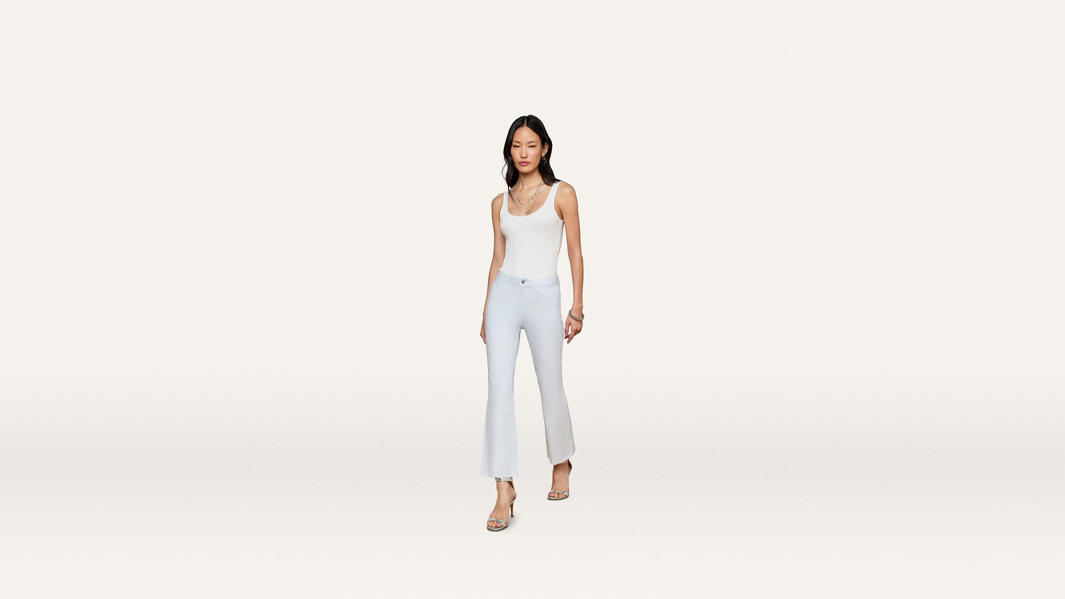 A woman in a white sleeveless top and light blue flared pants walks forward, wearing sandals and a delicate necklace, set against a plain white background.