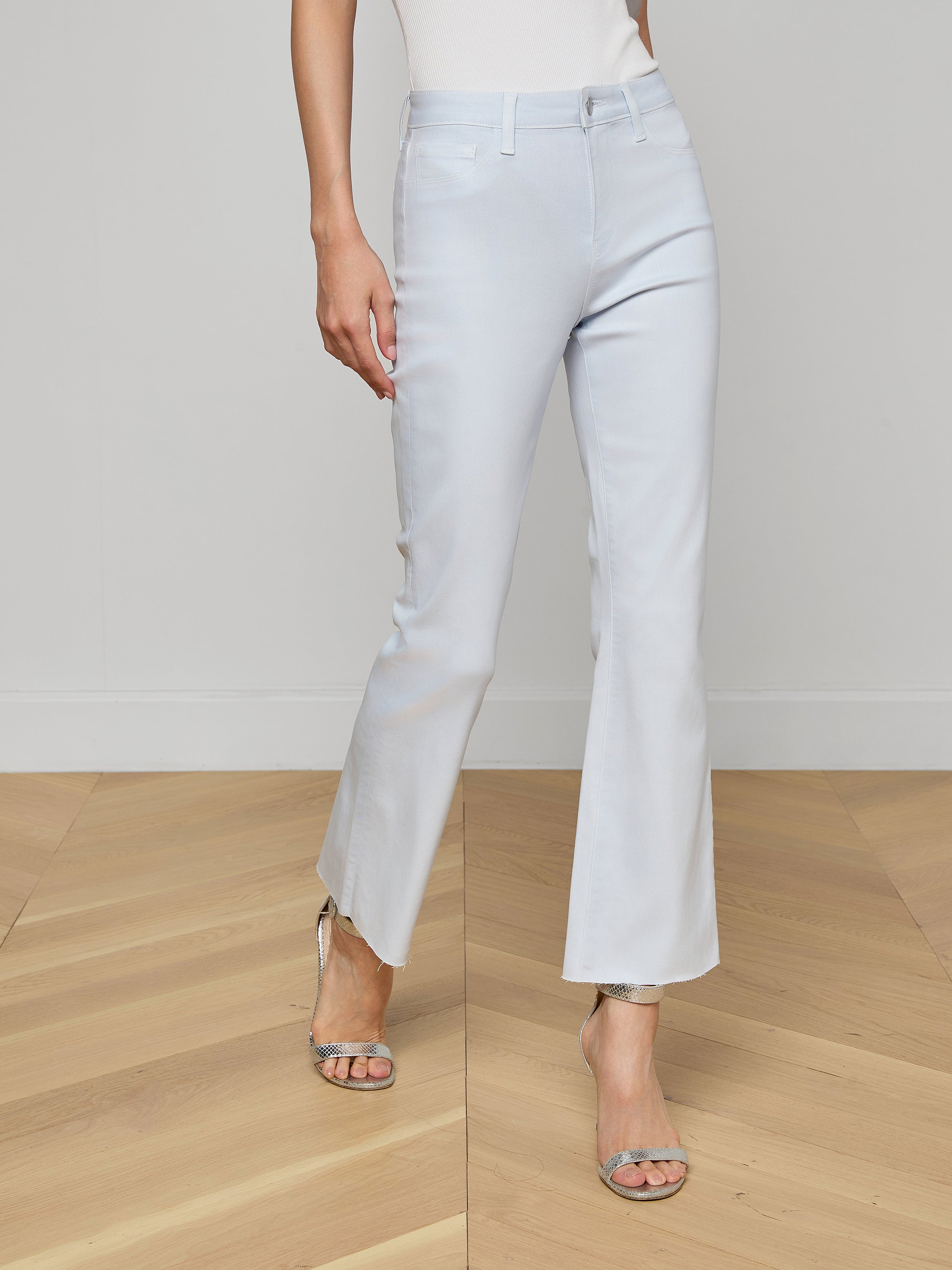 Someone wearing the Kendra Coated Cropped Flare Jean in light blue with silver heeled sandals stands on a wooden floor against a plain white wall, showing only the lower half of their body.