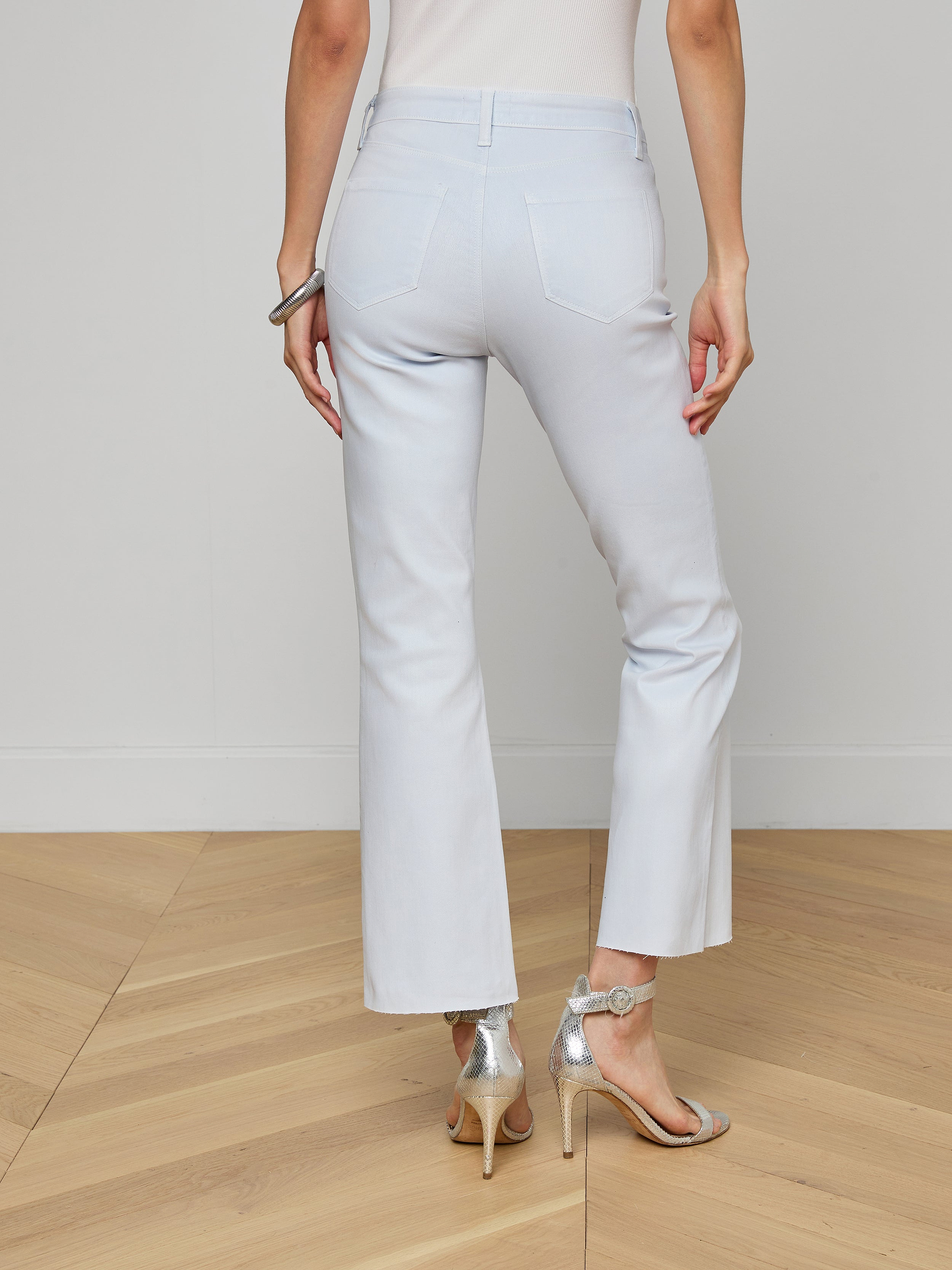 A person stands on a wood floor facing away, wearing the Kendra Coated Cropped Flare Jean in light blue, paired with a white top, silver bracelet, and silver high-heeled sandals. The background features a plain light-colored wall.
