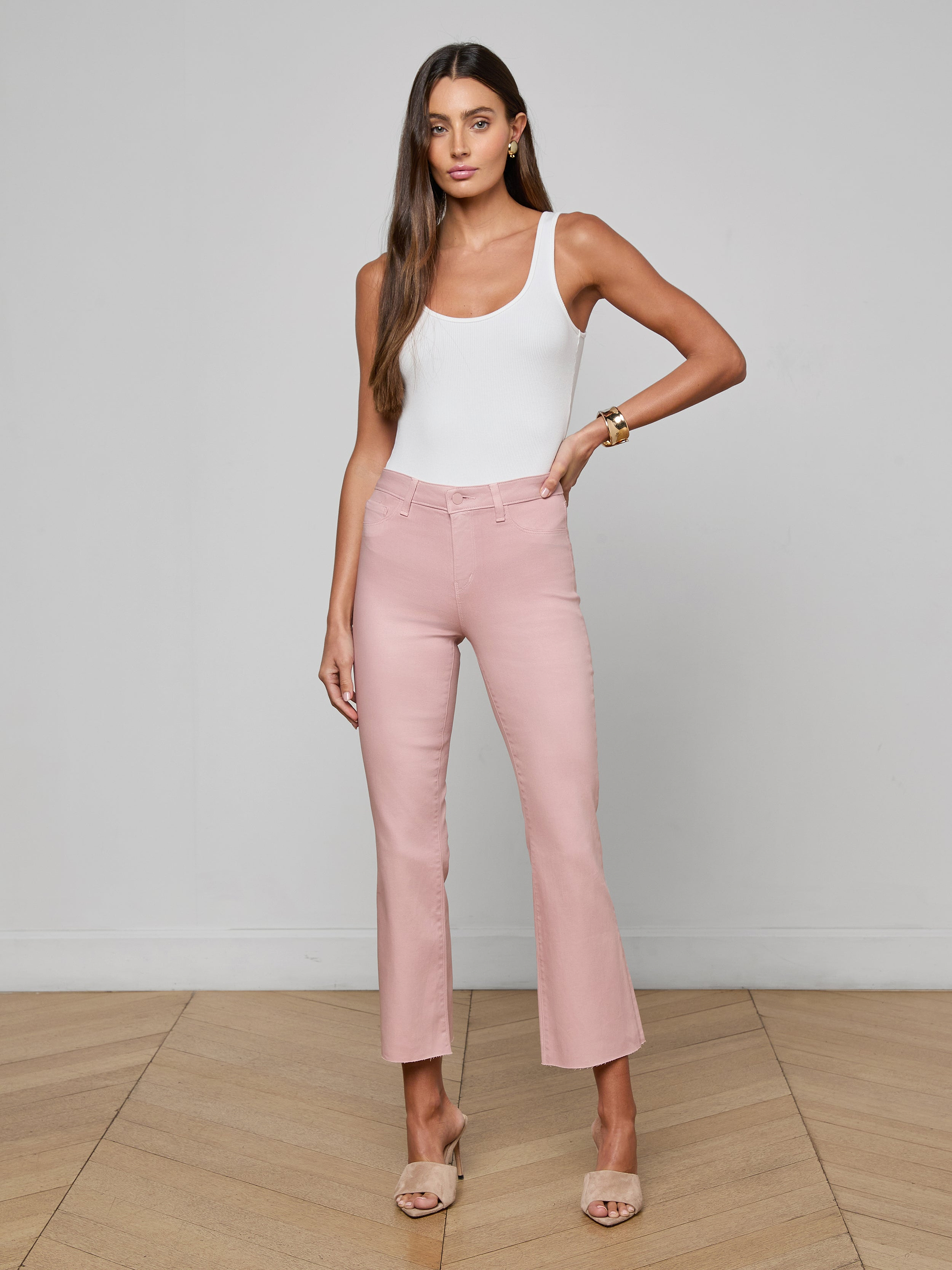 A woman stands against a plain white background wearing a white tank top, the Kendra Coated Cropped Flare Jean in pink—a high-waisted, leather-alternative coated denim—and beige open-toe heels. She has long hair and a neutral expression.