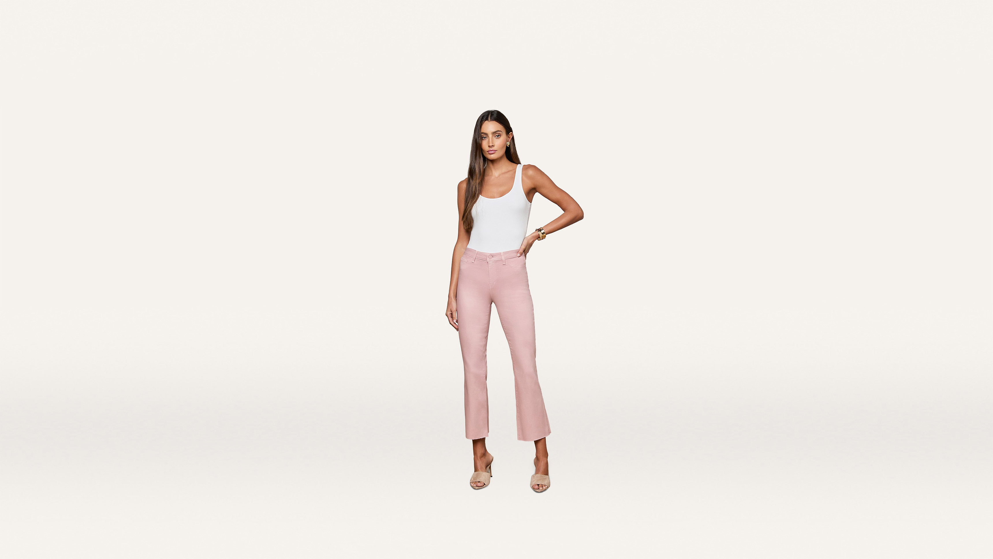 A woman stands against a white background, wearing a white sleeveless top paired with pink high-waisted cropped pants and nude heels. She has long hair and her left hand rests confidently on her hip, creating a chic, modern look.
