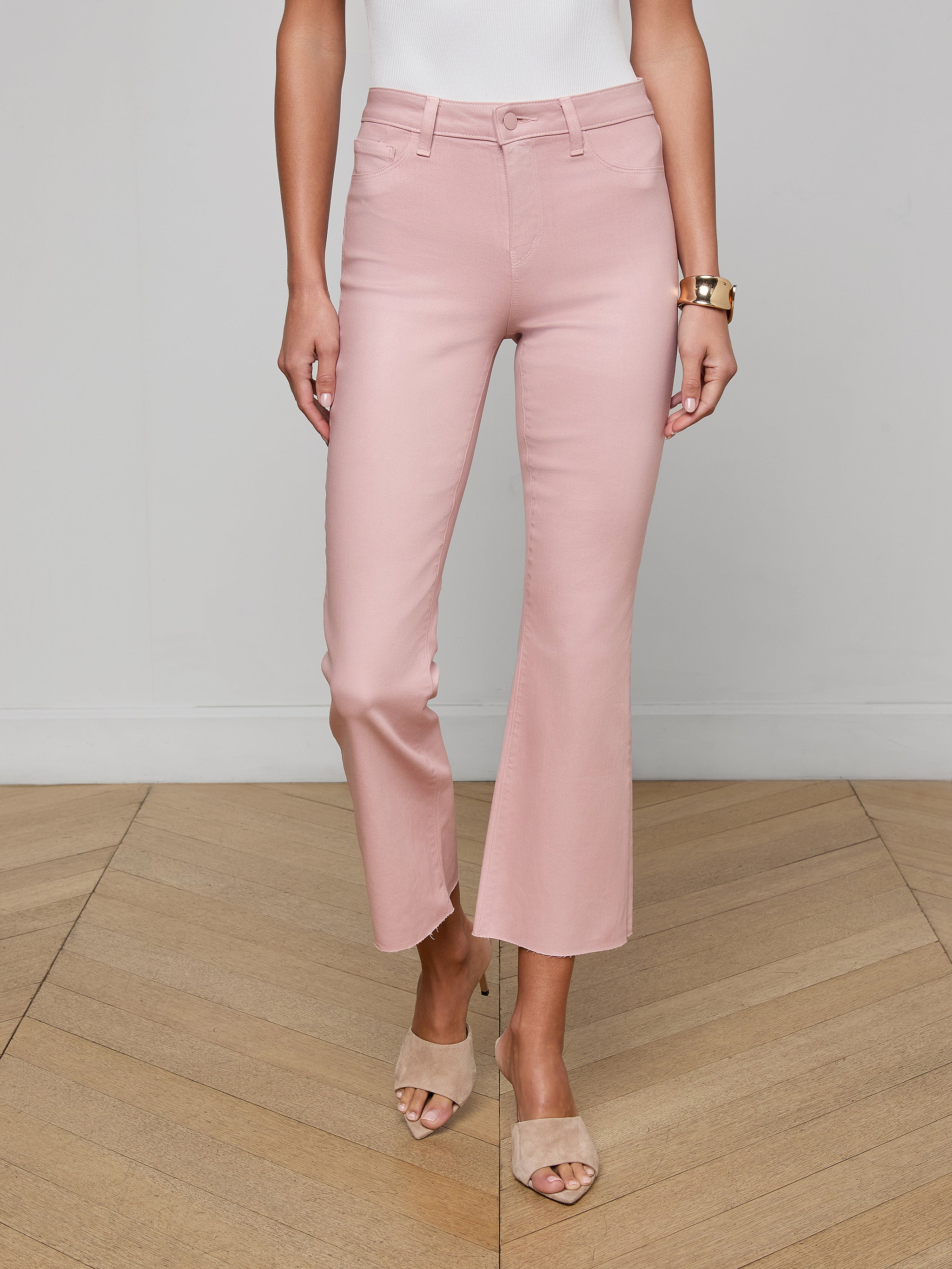 A woman wearing the Kendra Coated Cropped Flare Jean in blush pink, paired with a white sleeveless top, beige heeled sandals, and a gold watch, stands against a plain white background with only the lower half of her body visible.