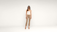 Woman wearing sunglasses, a white tank top, tan pants, and heeled sandals stands against a plain white background, facing forward with her arms relaxed by her sides.