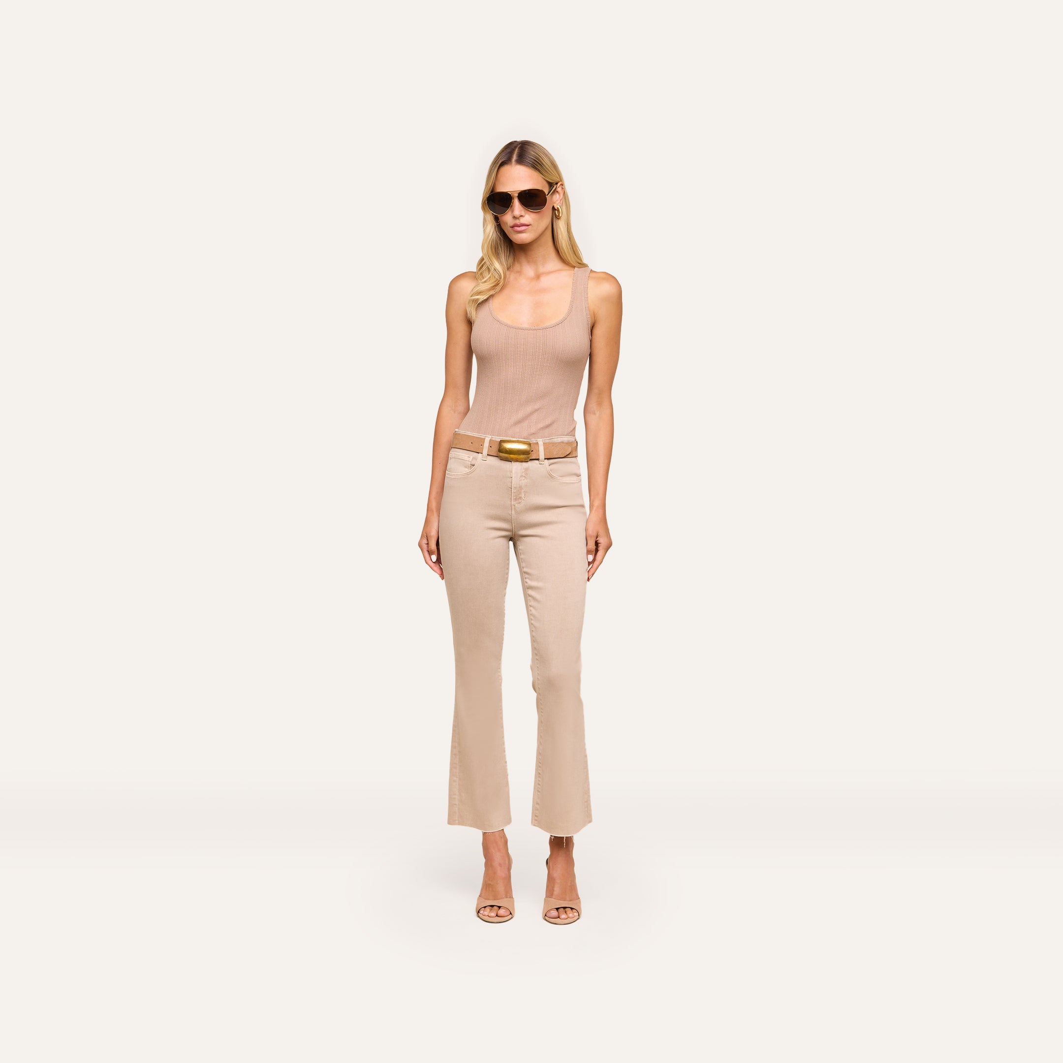 Woman wearing sunglasses, a beige sleeveless top, Kendra Cropped Flare Jean with a belt, and beige heels stands against a plain white background.