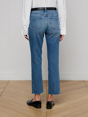 A person stands indoors on a wooden floor, facing away, wearing a white long-sleeve shirt, a black belt, black shoes, and L'AGENCE Milana Slouchy Stovepipe Jean—high-waisted vintage wash all-cotton denim with frayed hems—against a plain white wall.