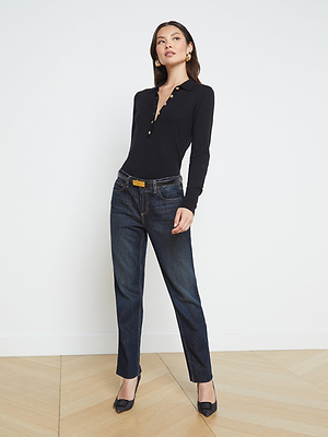 A woman stands in a minimal room wearing a fitted black long-sleeve top with gold buttons, dark blue jeans, black heels, and gold earrings, posing confidently with one hand in her pocket.
