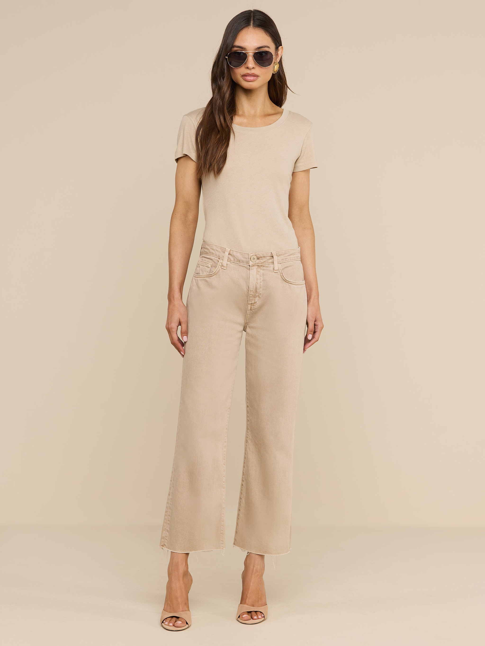 Una donna dai lunghi capelli castani, con indosso occhiali da sole, una maglietta a maniche corte color beige neutro, i jeans Wanda Cropped Wide-Leg e tacchi color carne, è in posa davanti a uno sfondo beige a tinta unita.