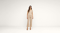 A woman wearing sunglasses, a beige t-shirt, wide-legged beige pants, and heeled sandals stands against a plain white background.