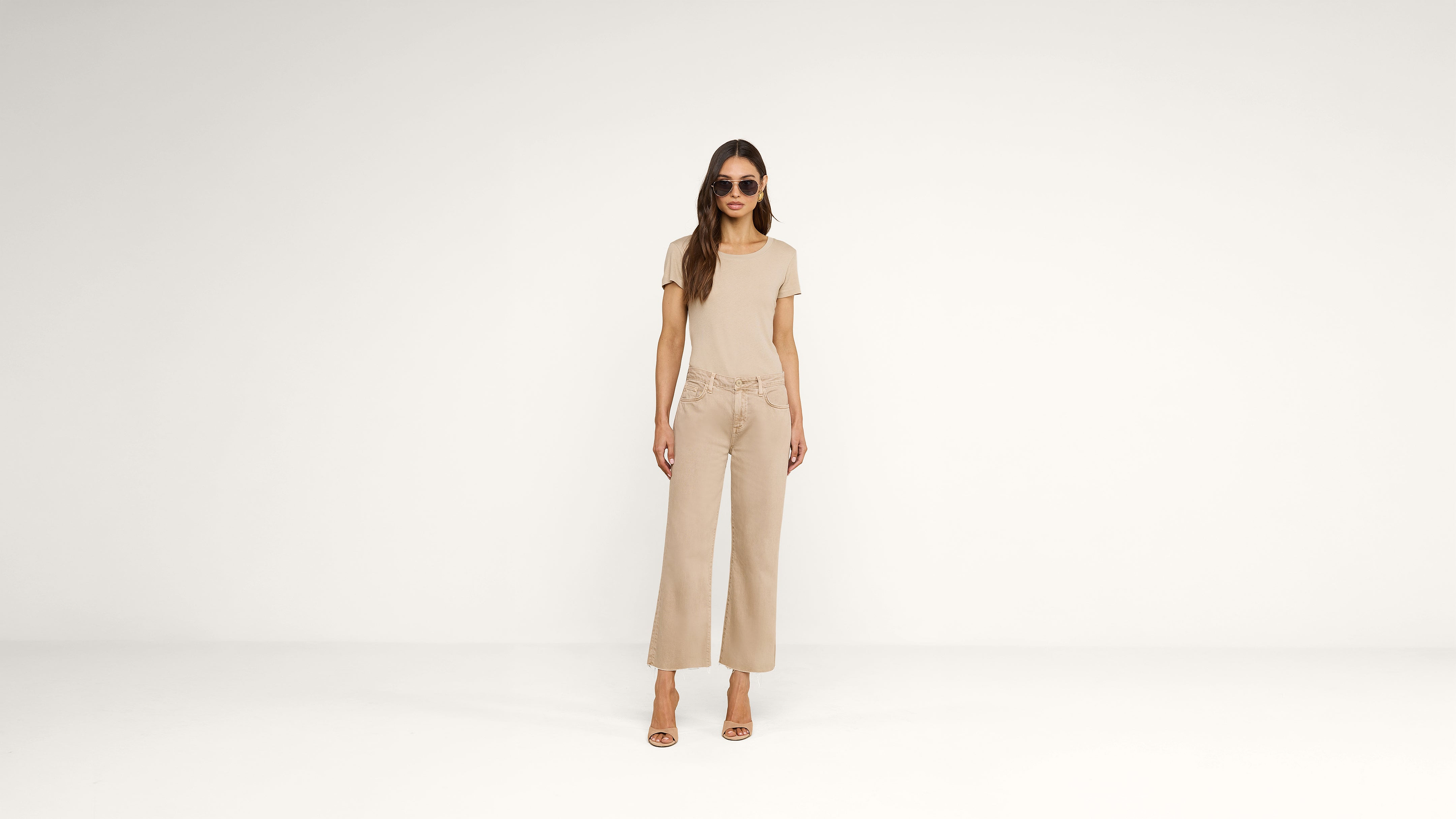 A woman wearing sunglasses, a beige t-shirt, wide-legged beige pants, and heeled sandals stands against a plain white background.