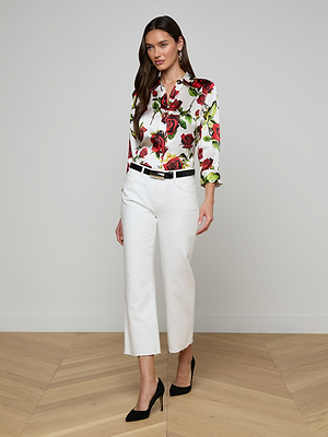 A woman stands on a wooden floor wearing a floral blouse, the Wanda Cropped Wide-Leg Jean in white, a black belt, and black heels against a plain light-colored background.