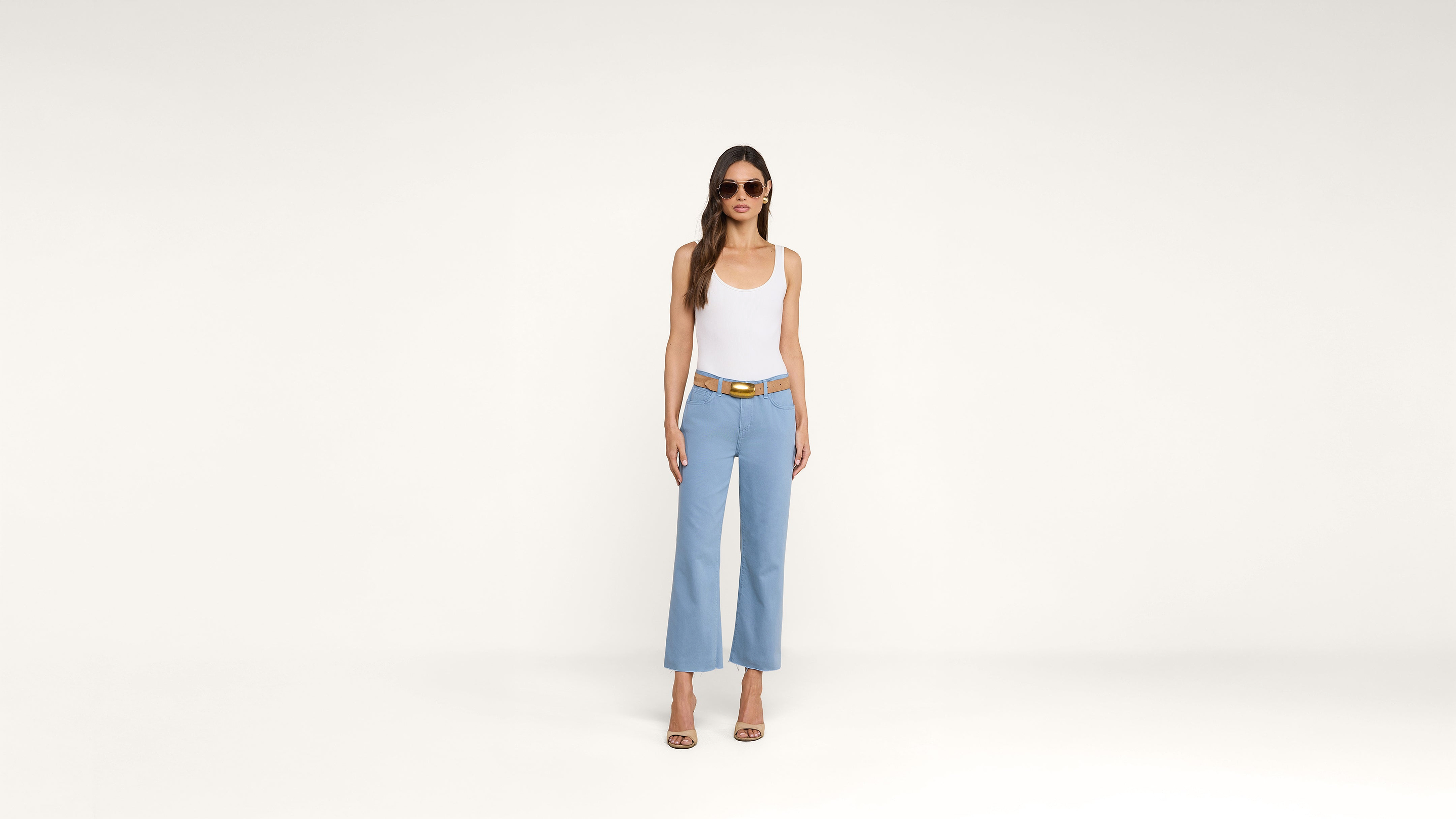 Woman wearing sunglasses, a white tank top, light blue wide-leg pants, a tan belt, and heels stands against a plain white background.