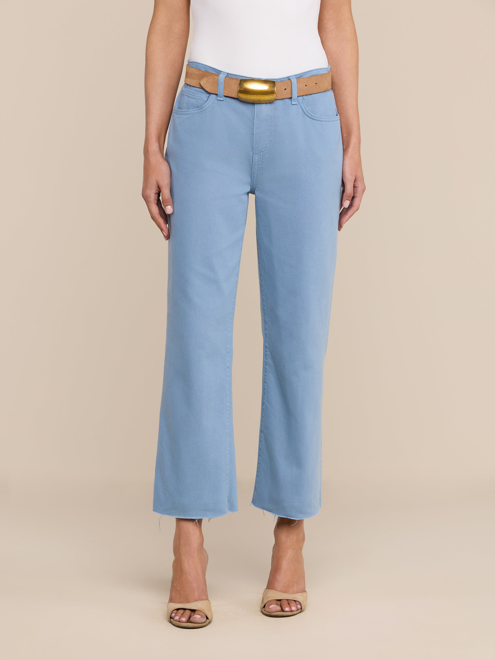 Une personne porte le jean court à jambes larges « Wanda » bleu clair à ourlet brut, associé à une ceinture beige clair, un haut blanc et des escarpins beiges à bout ouvert, le tout sur un fond beige uni, pour un look parisien tout en simplicité.