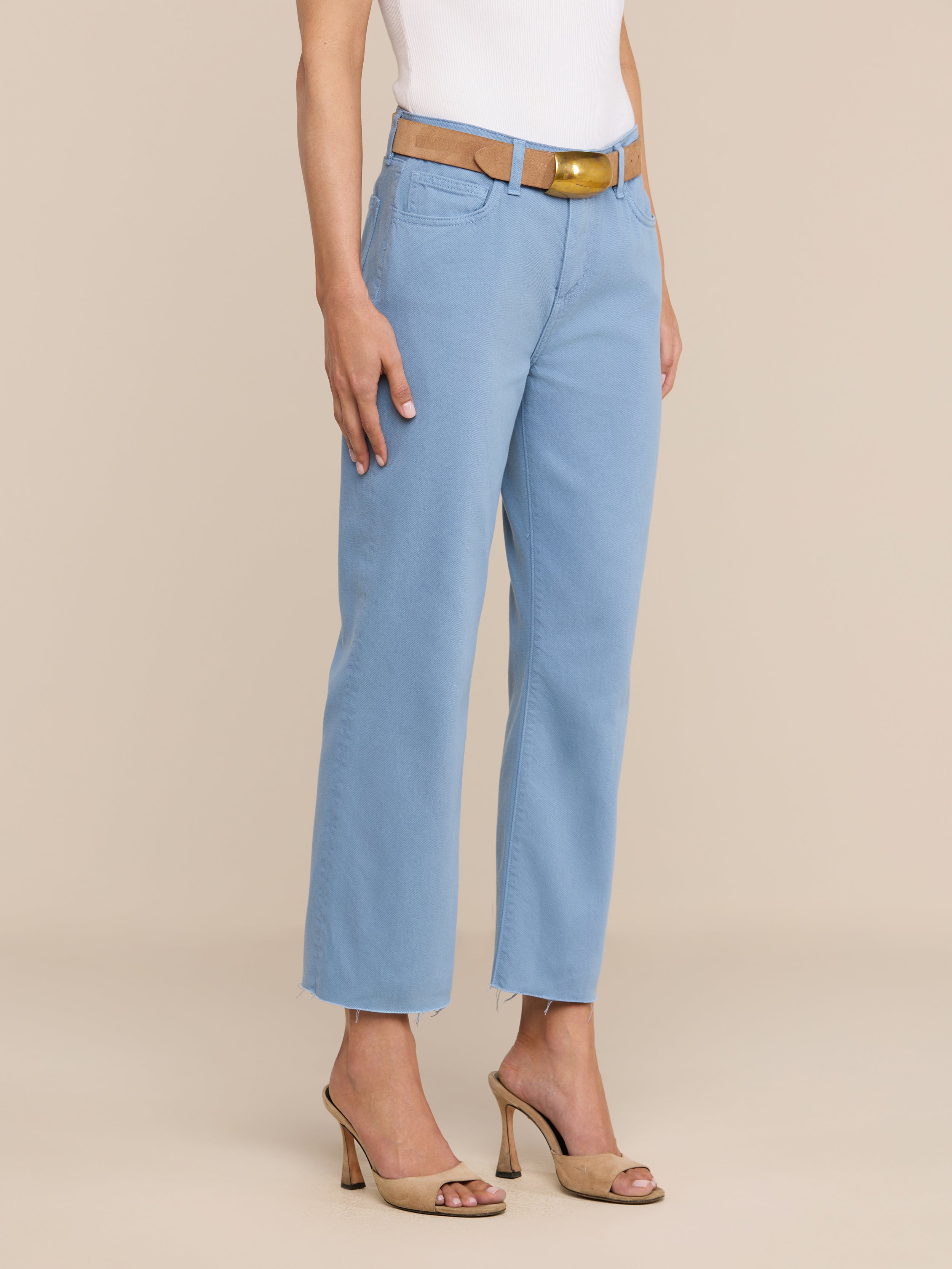 Une personne portant un jean court à jambes larges « Wanda » bleu clair, un haut sans manches blanc, une ceinture marron et des sandales à talons hauts beiges se tient debout devant un fond beige uni.