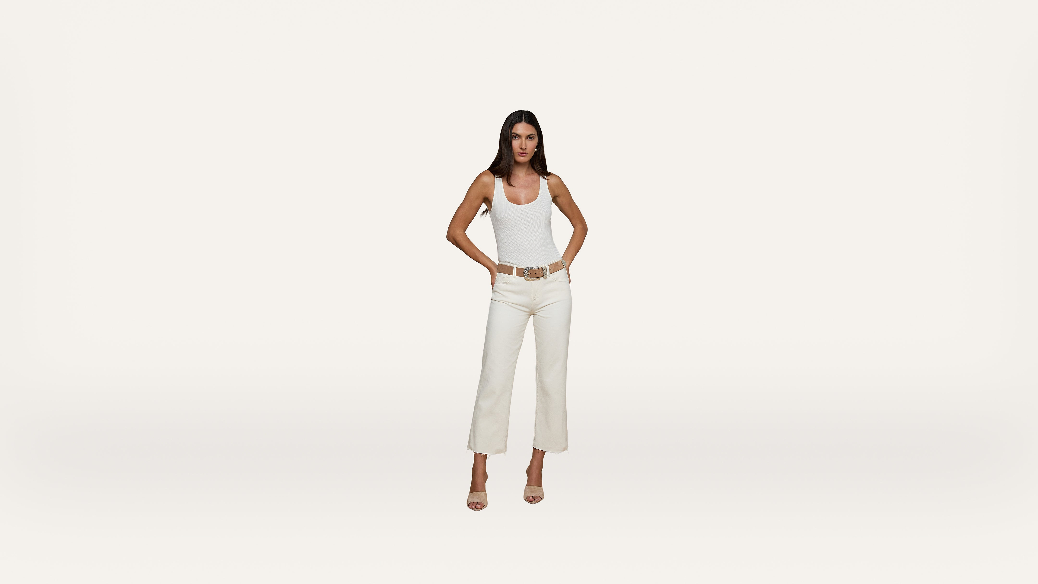 A woman stands facing forward with her hands on her hips, wearing a white tank top, white pants, and beige heels against a plain white background.