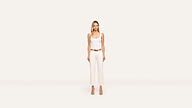 A woman stands facing forward, wearing a white sleeveless top, white high-waisted pants, a tan belt, and tan heels against a plain off-white background. The outfit highlights her elegant style.