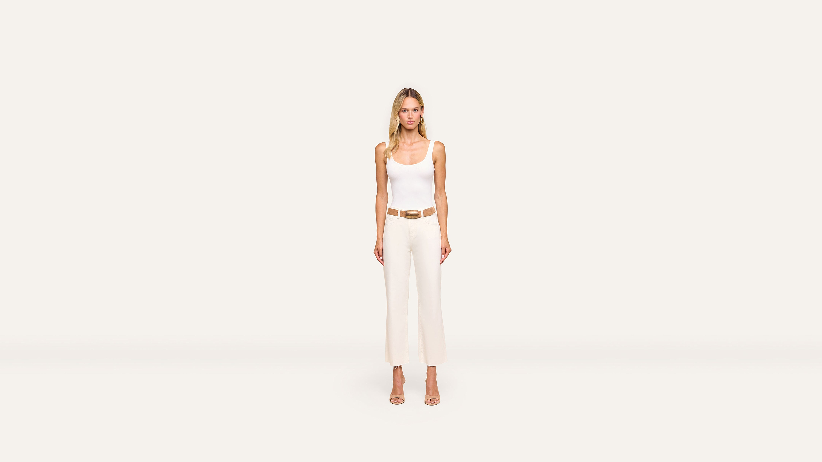A woman stands facing forward, wearing a white sleeveless top, white high-waisted pants, a tan belt, and tan heels against a plain off-white background. The outfit highlights her elegant style.