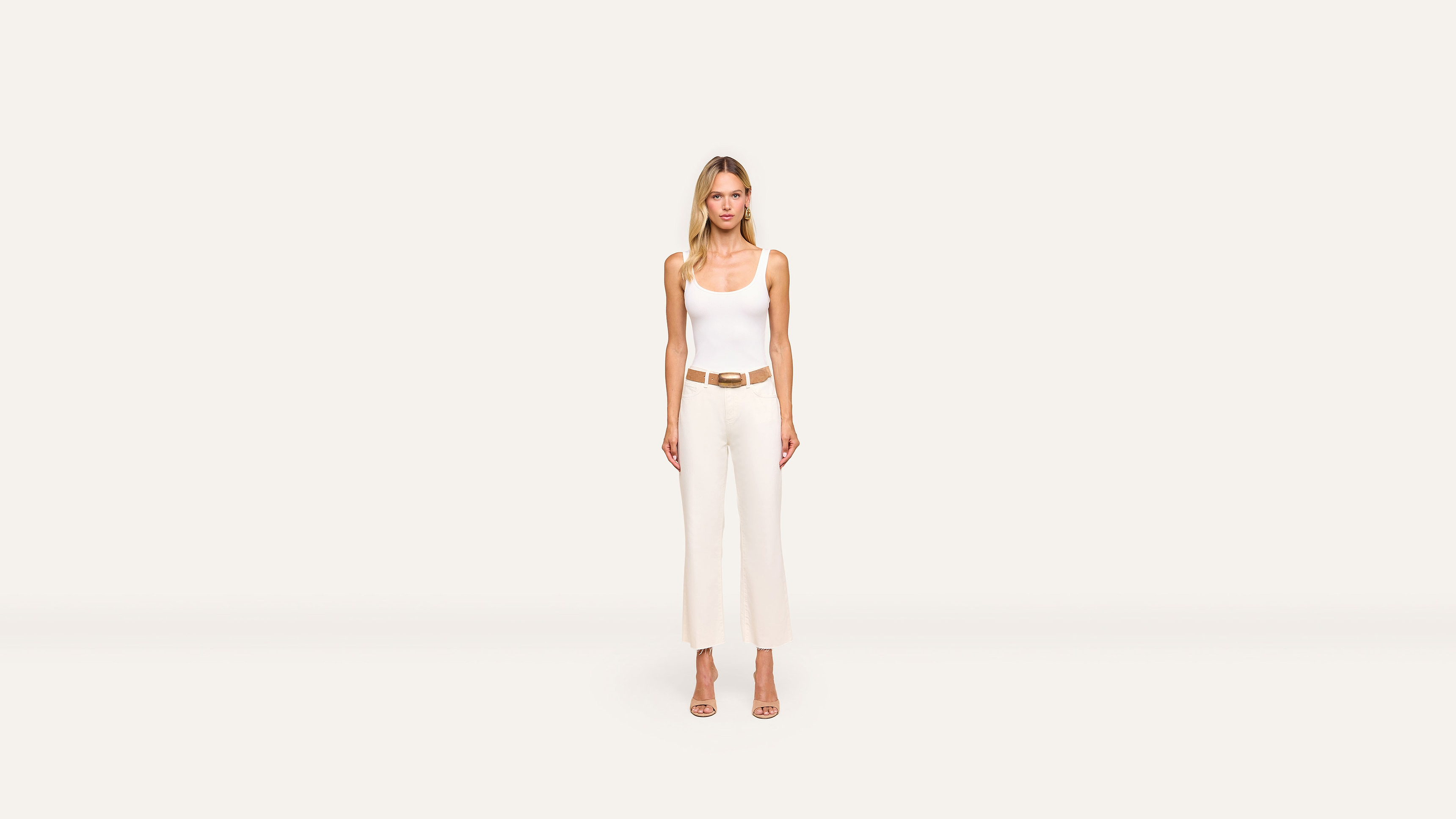 A woman stands facing forward, wearing a white sleeveless top, white high-waisted pants, a tan belt, and tan heels against a plain off-white background. The outfit highlights her elegant style.