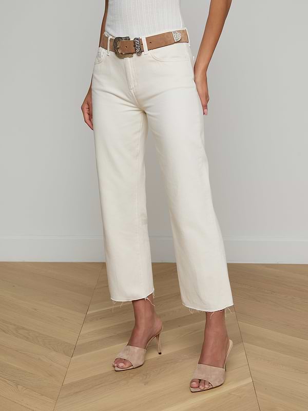 A woman models the Wanda Cropped Wide-Leg Jean in off-white, styled with a brown belt, a white ribbed sleeveless top, and beige heeled sandals. The image is set against a plain light background, and the woman’s head is not visible.