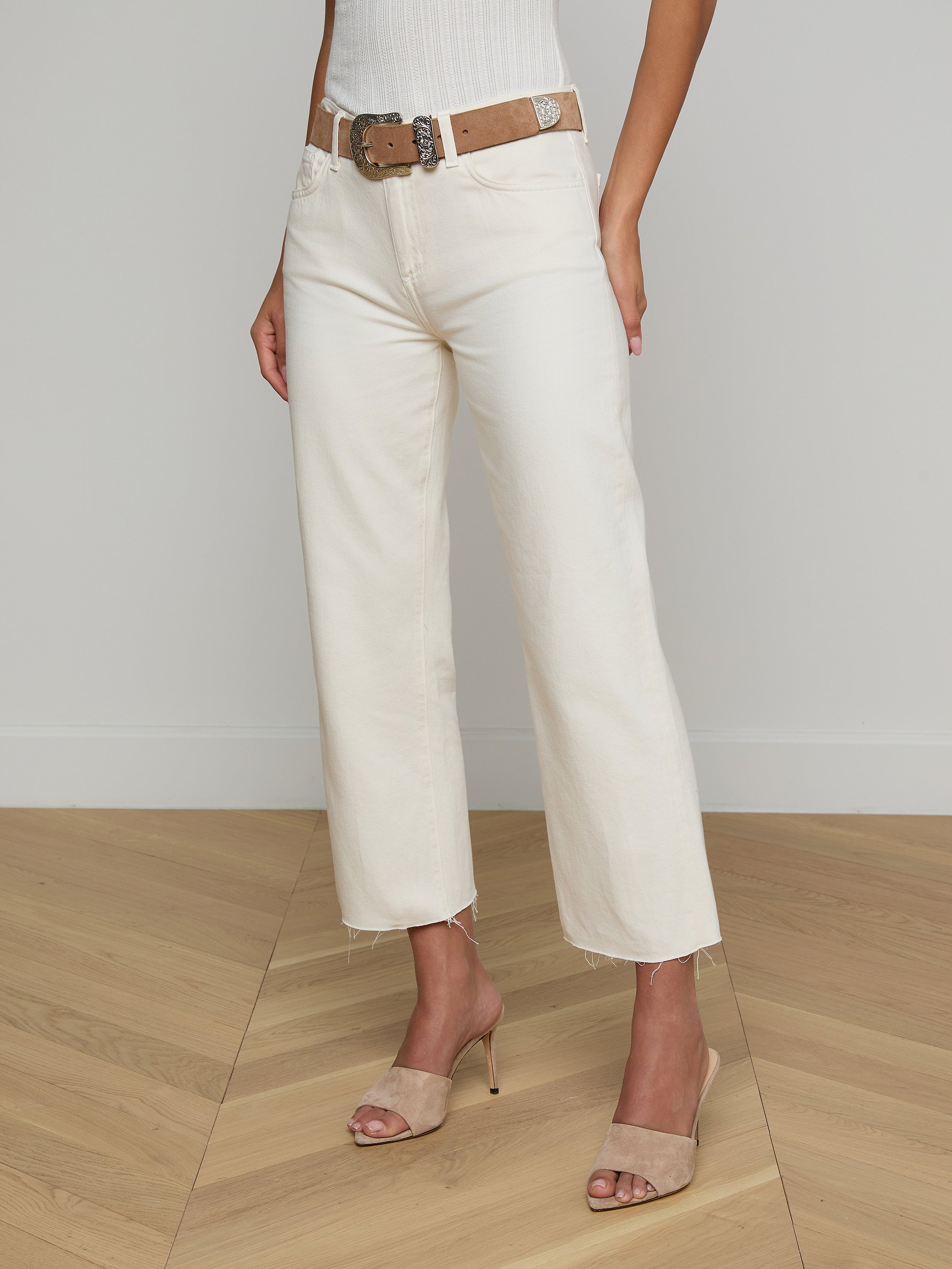 A woman models the Wanda Cropped Wide-Leg Jean in off-white, styled with a brown belt, a white ribbed sleeveless top, and beige heeled sandals. The image is set against a plain light background, and the woman’s head is not visible.
