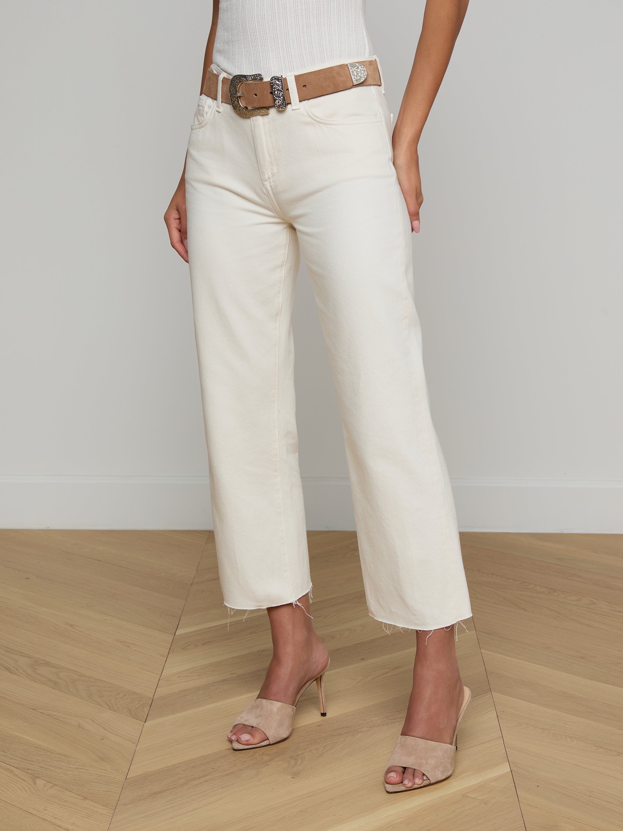 A woman models the Wanda Cropped Wide-Leg Jean in off-white, styled with a brown belt, a white ribbed sleeveless top, and beige heeled sandals. The image is set against a plain light background, and the woman’s head is not visible.