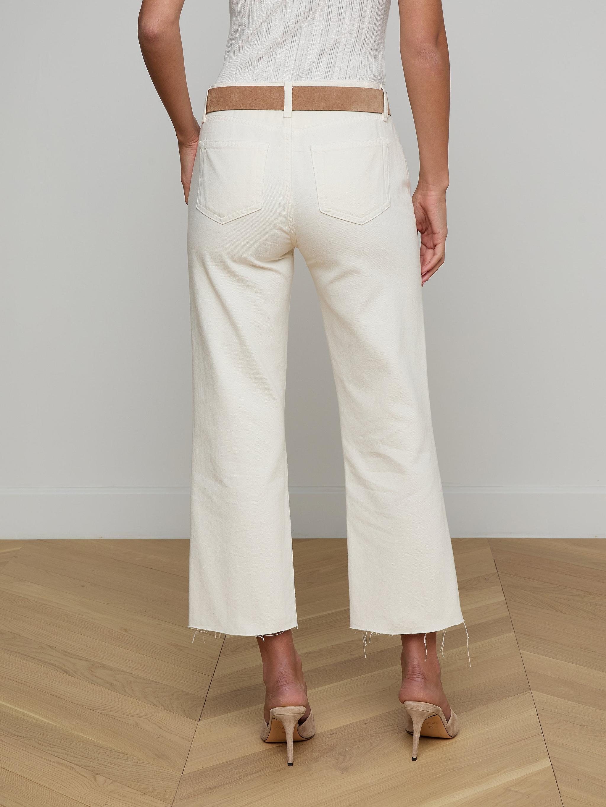 Wearing the Wanda Cropped Wide-Leg Jean in an off-white ecru wash with frayed hems, a woman stands facing away, styled with a beige belt and beige high-heeled shoes against a plain white background.