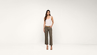 A woman stands against a plain white background, wearing a white tank top, olive green pants, a belt, and heeled sandals. She has long brown hair and a neutral expression.