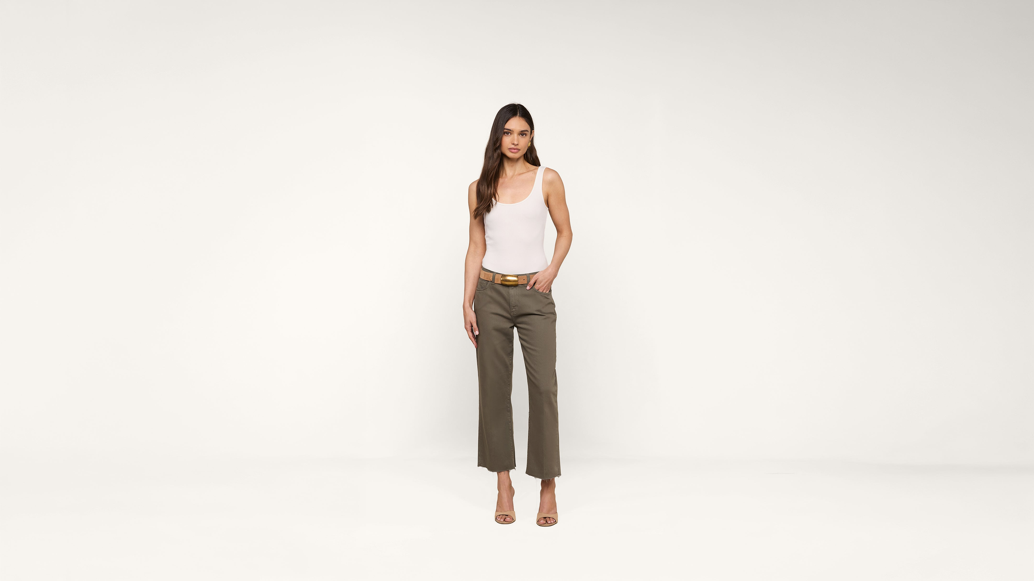 A woman stands against a plain white background, wearing a white tank top, olive green pants, a belt, and heeled sandals. She has long brown hair and a neutral expression.