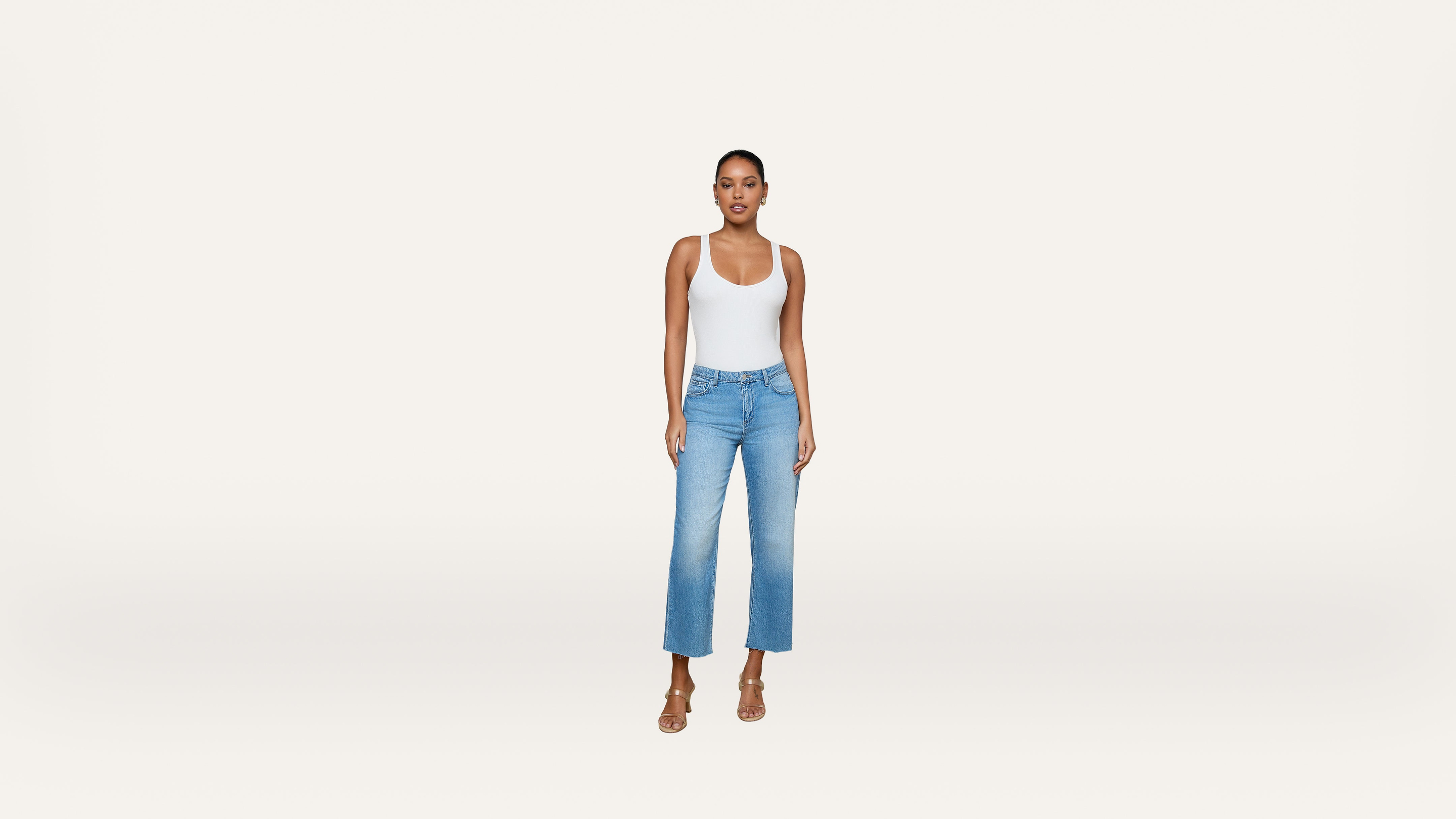 A person stands facing forward, wearing a white tank top, light blue high-waisted jeans, and beige heeled sandals against a plain white background.