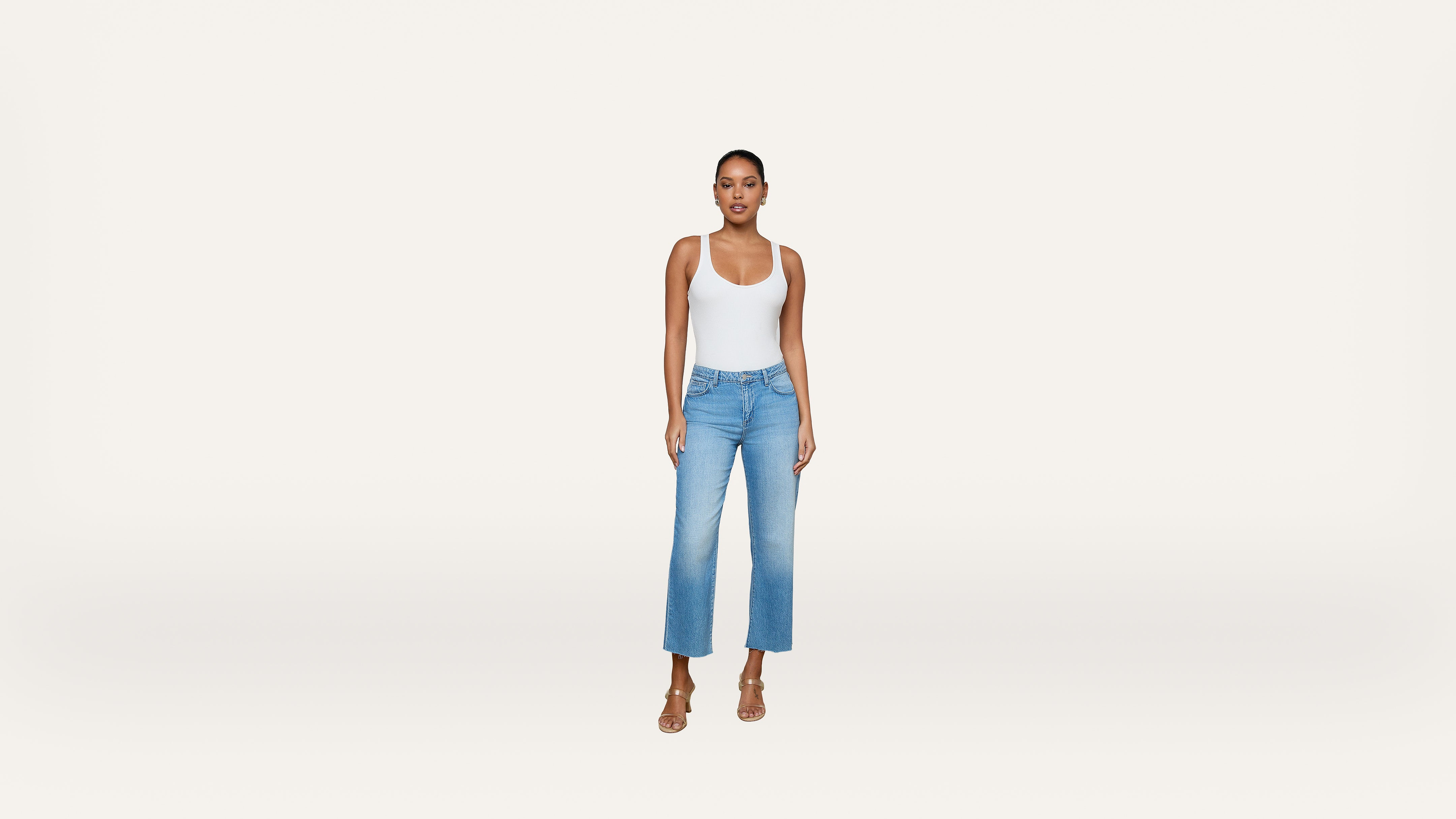 A person stands facing forward, wearing a white tank top, light blue high-waisted jeans, and beige heeled sandals against a plain white background.