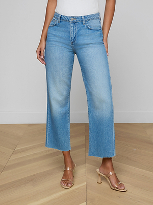 Wearing the Wanda Cropped Wide-Leg Jean in light blue with a raw hem, styled Parisian-chic with a white top and clear heeled sandals, a person stands on a light wood floor against a plain white wall.