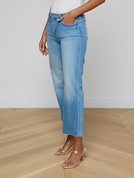 A woman stands against a plain light background, wearing the Wanda Cropped Wide-Leg Jean—light blue, high-waisted, all-cotton denim jeans featuring raw hems and a cropped length—paired with a white top and tan heeled sandals.