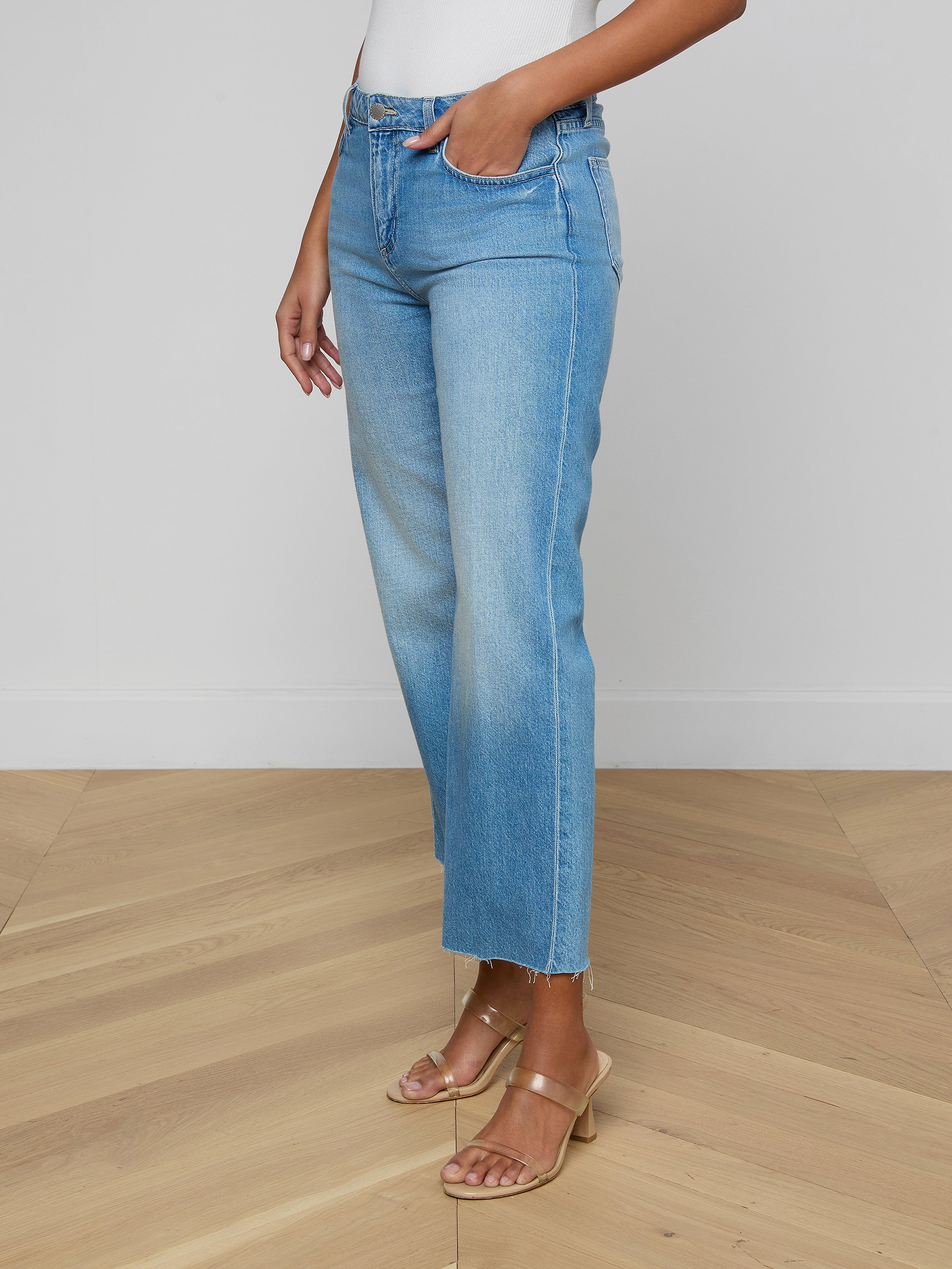 A woman stands against a plain light background, wearing the Wanda Cropped Wide-Leg Jean—light blue, high-waisted, all-cotton denim jeans featuring raw hems and a cropped length—paired with a white top and tan heeled sandals.