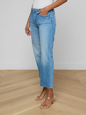 The lower half of a person is shown wearing Wanda Cropped Wide-Leg Jean in high-waisted light blue denim with a raw hem and a white top, standing on light wood flooring for an effortlessly Parisian look.