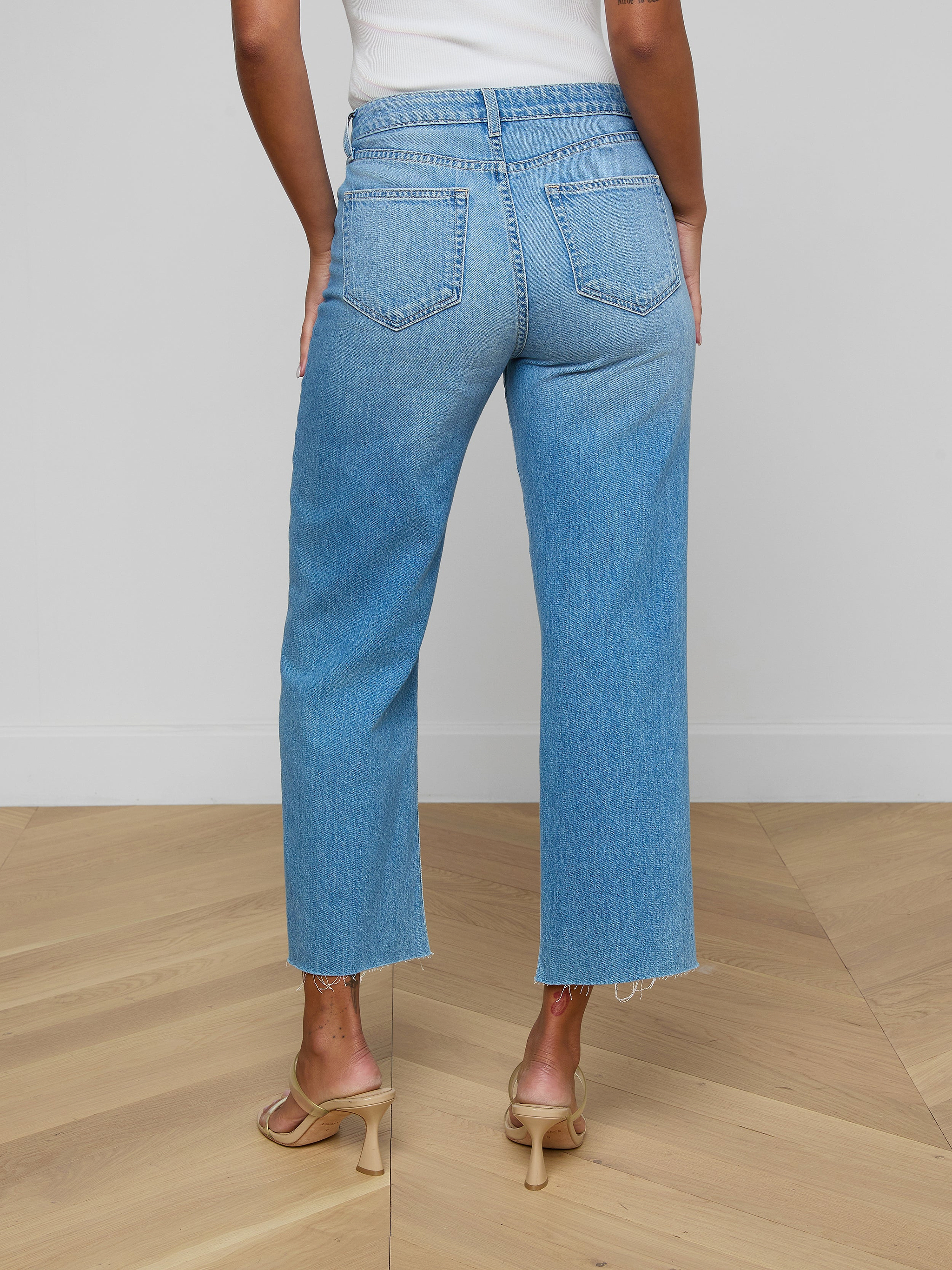 A person is seen from behind, standing against a plain white background, wearing the Wanda Cropped Wide-Leg Jean—light blue, high-waisted jeans crafted from 100 cotton denim—and beige heeled sandals.