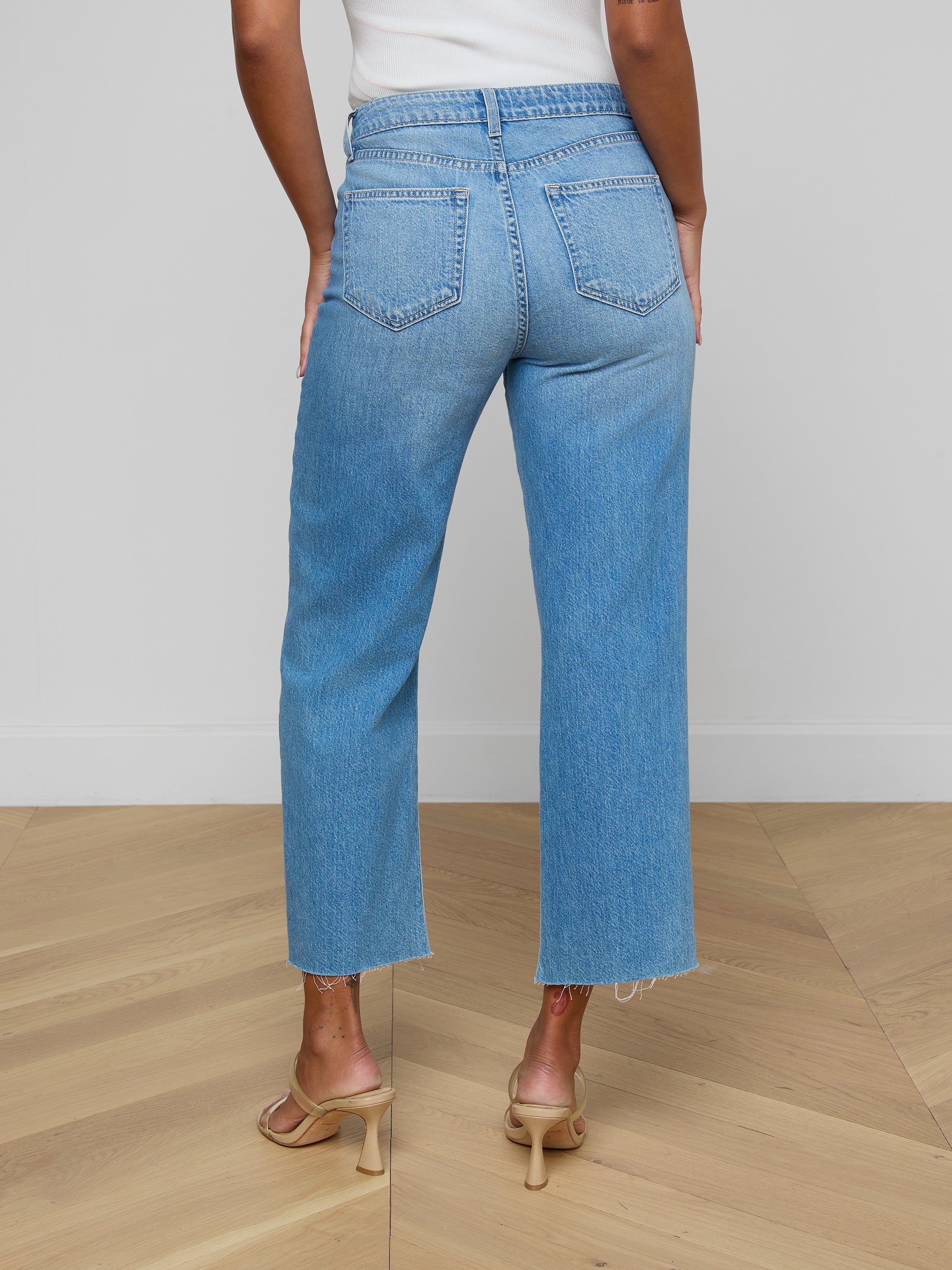 A person is seen from behind, standing against a plain white background, wearing the Wanda Cropped Wide-Leg Jean—light blue, high-waisted jeans crafted from 100 cotton denim—and beige heeled sandals.