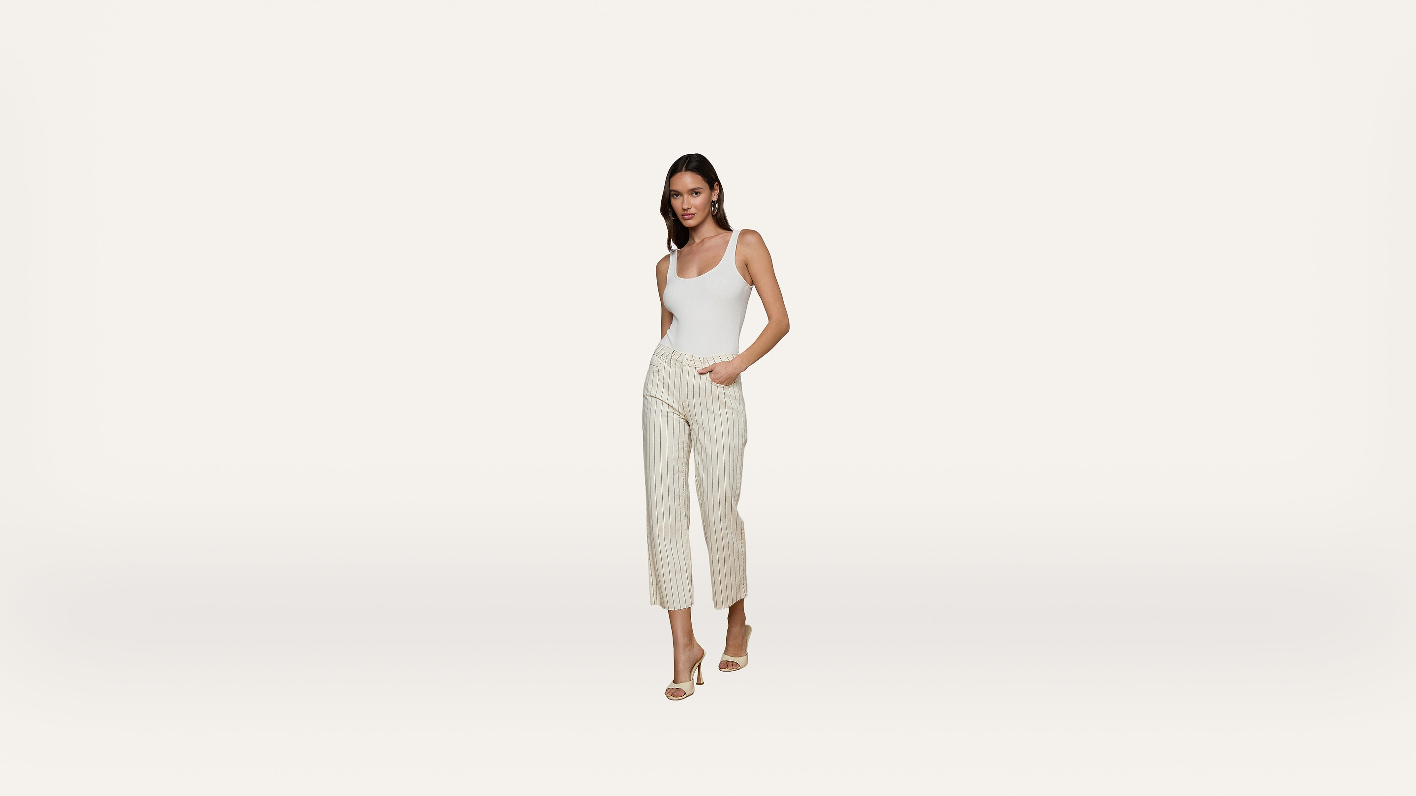 Woman wearing a white sleeveless top and light-colored, striped pants with beige high-heeled sandals, standing against a plain white background.