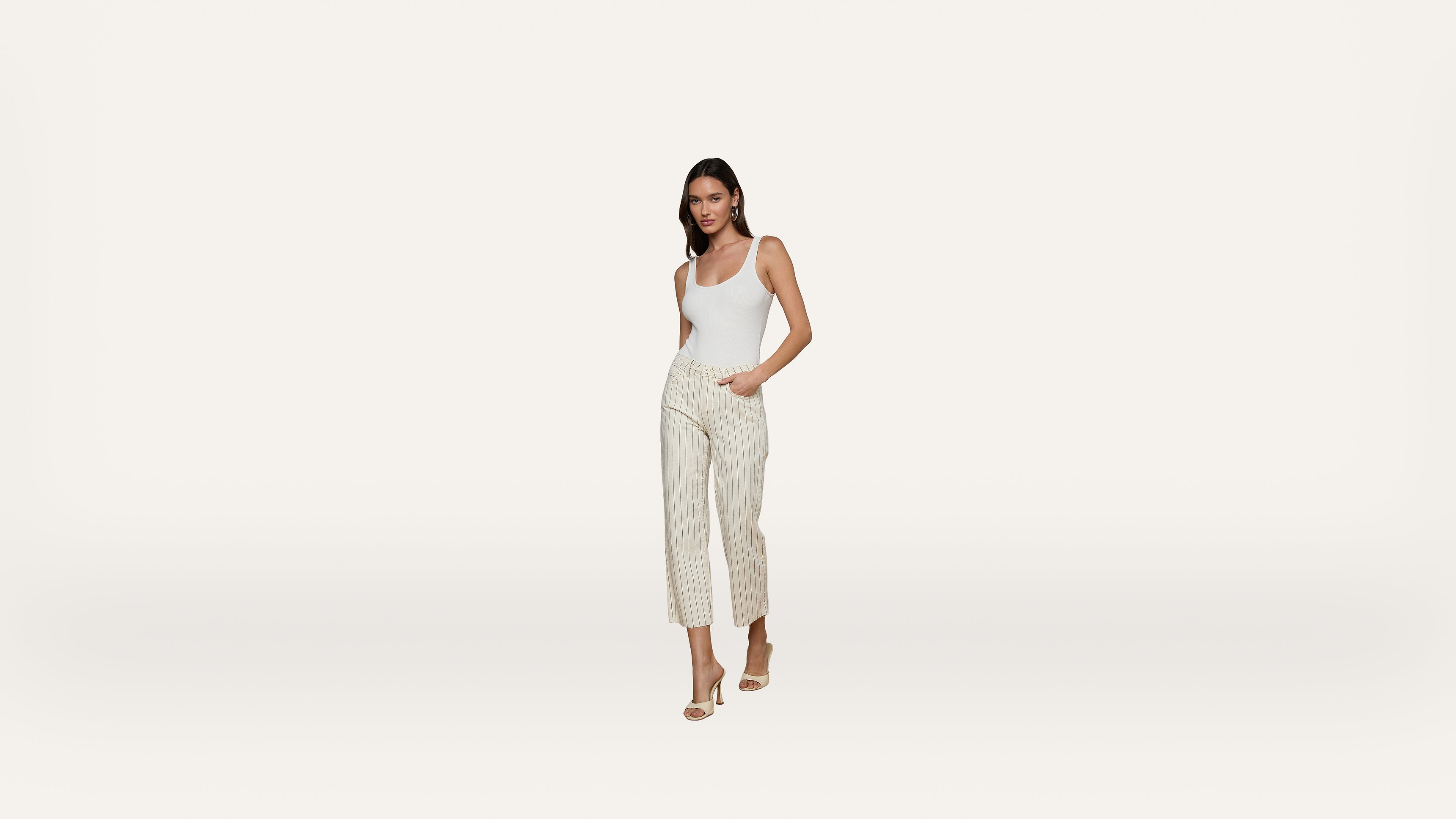 Woman wearing a white sleeveless top and light-colored, striped pants with beige high-heeled sandals, standing against a plain white background.