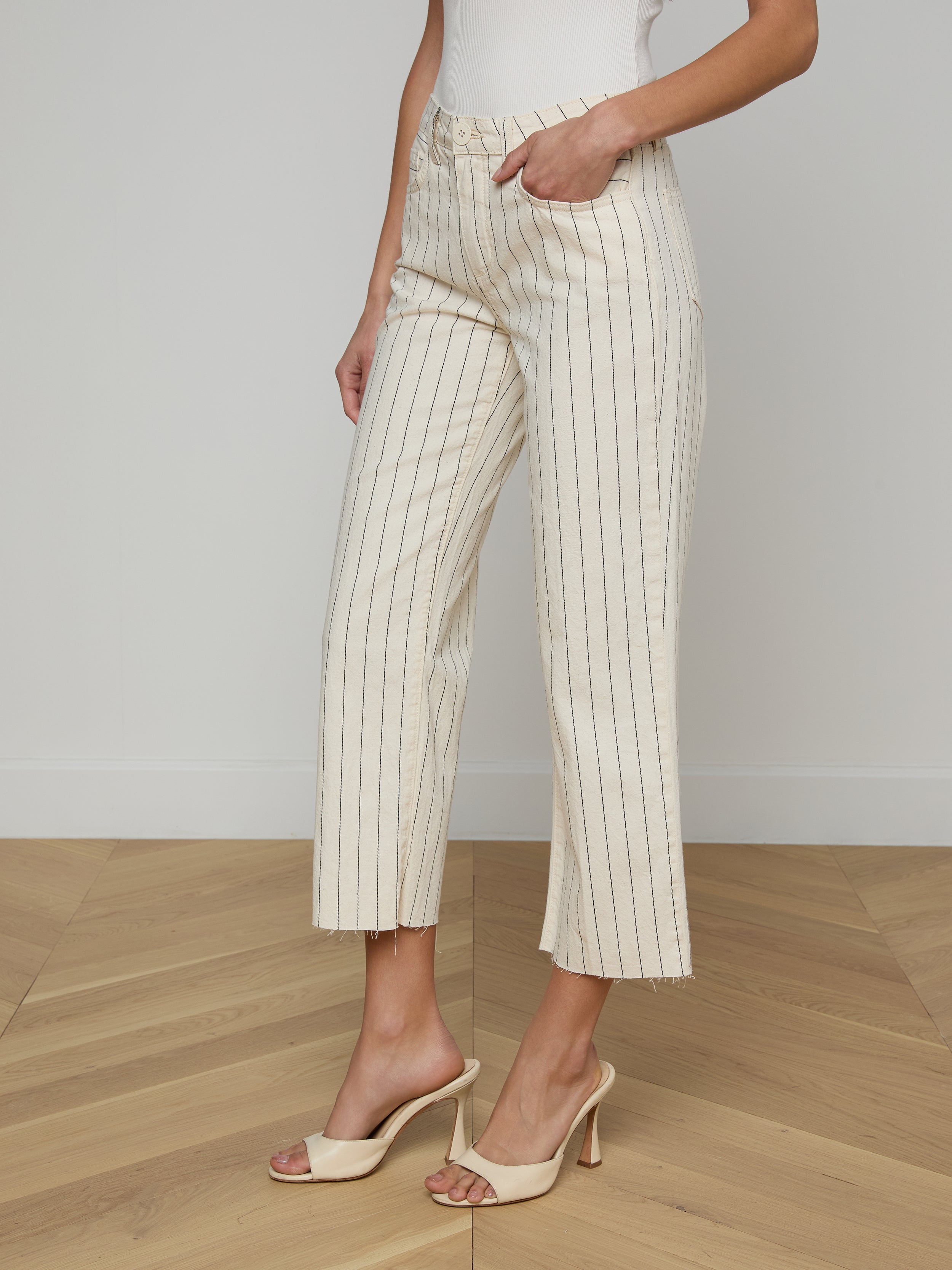 A woman wearing a white sleeveless top, the Wanda Cropped Wide-Leg Pant in cream-colored cotton twill with nautical stripes, and beige high-heeled sandals stands against a light background.