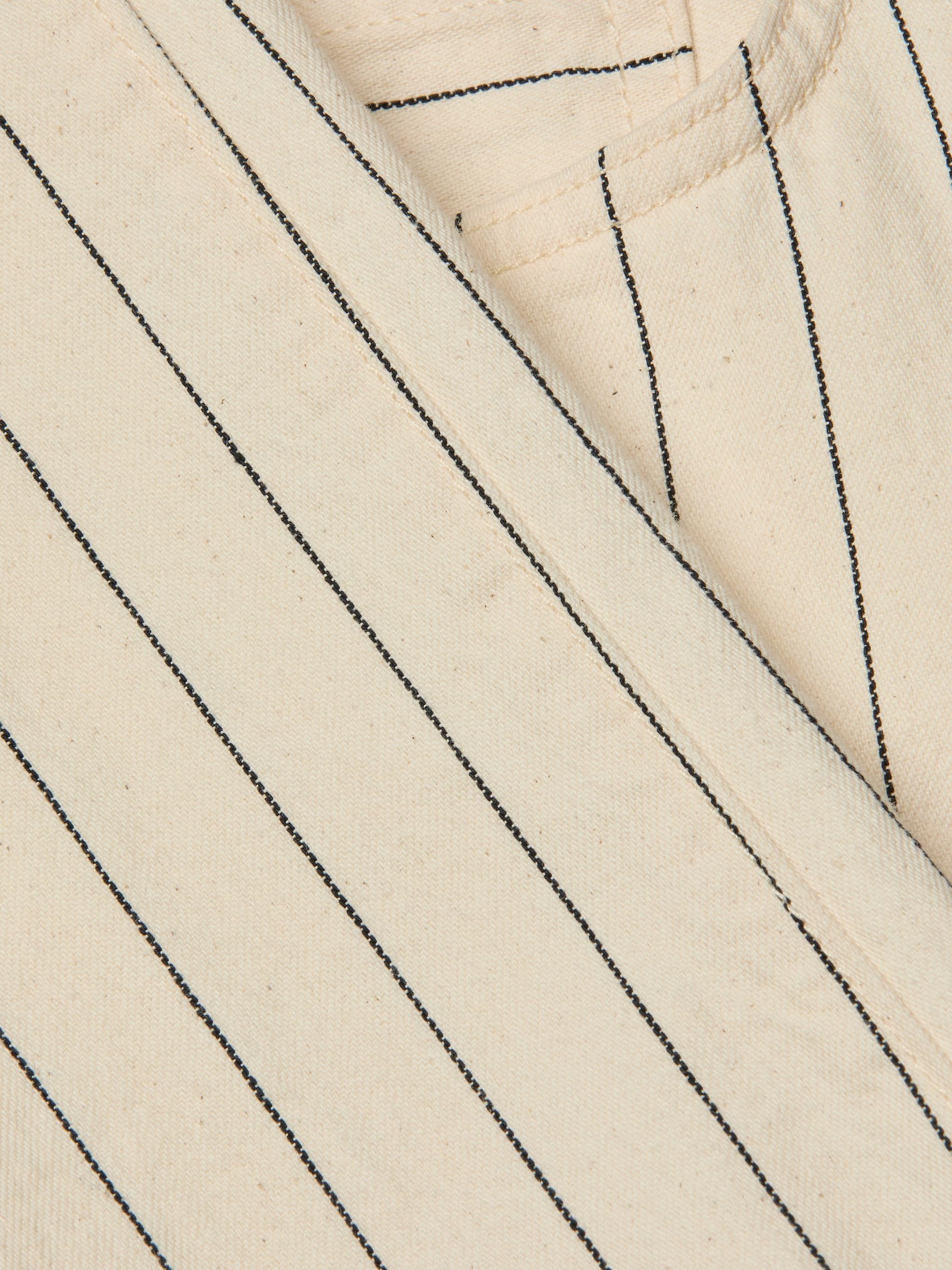Close-up of the Wanda Cropped Wide-Leg Pant in beige cotton twill with black vertical pinstripes in a nautical stripe pattern, showing part of a seam and pocket. The texture of the fabric and stitching details are visible.