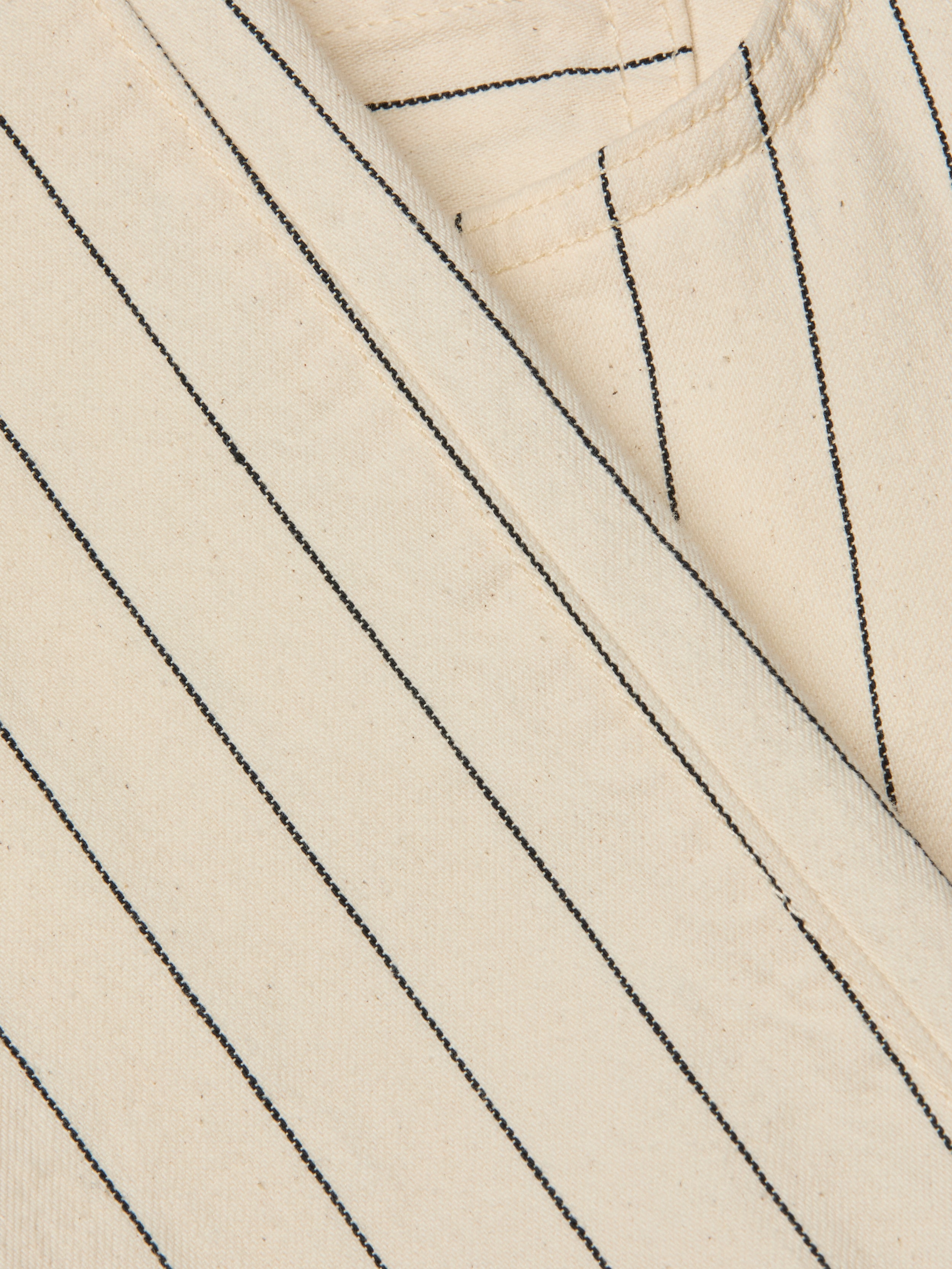 Close-up of the Wanda Cropped Wide-Leg Pant in beige cotton twill with black vertical pinstripes in a nautical stripe pattern, showing part of a seam and pocket. The texture of the fabric and stitching details are visible.