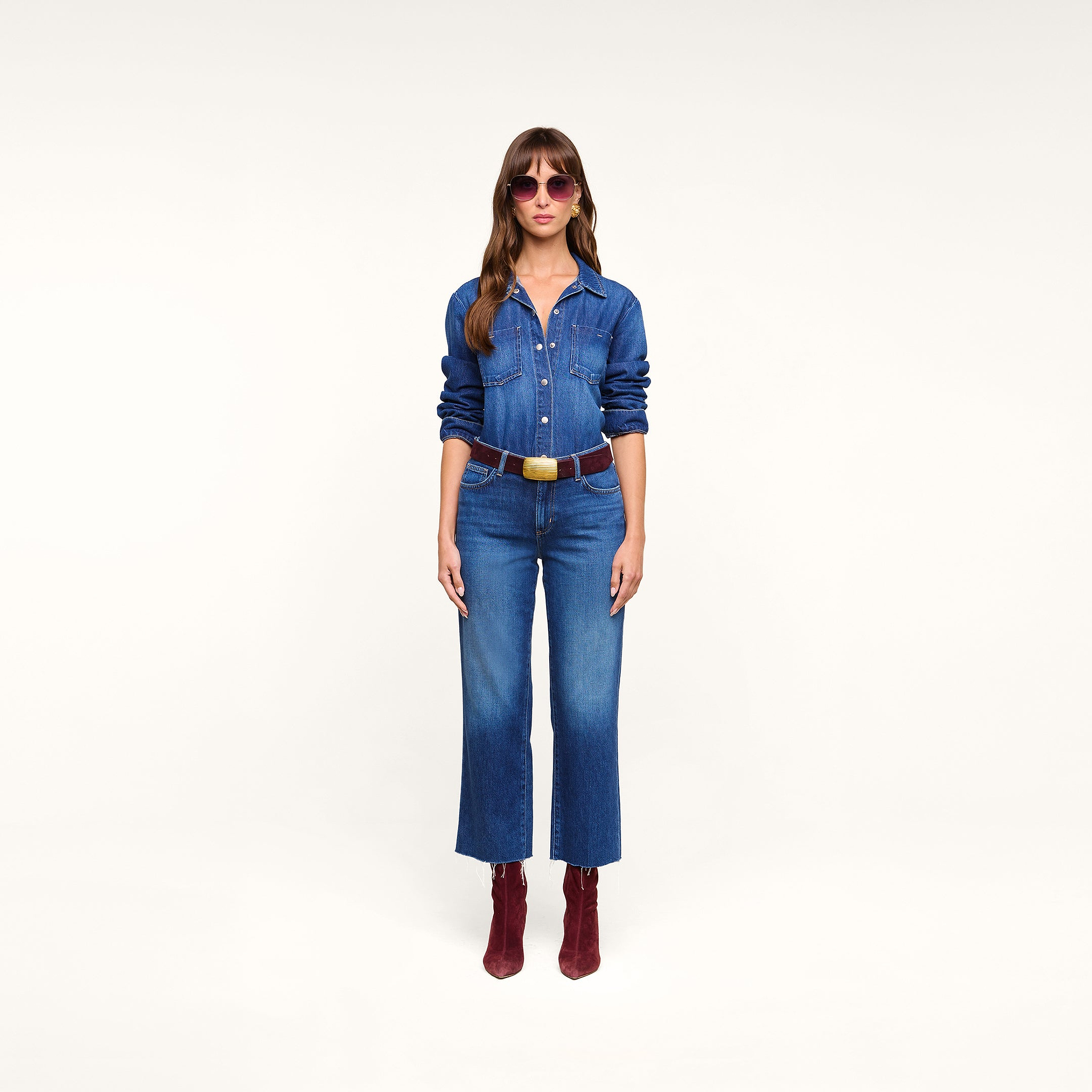 A woman stands facing forward, wearing sunglasses, a blue featherweight denim shirt, the Wanda Cropped Wide-Leg Jean, a brown belt, and red ankle boots, set against a plain white background.