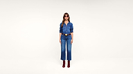 Woman wearing pink sunglasses, a denim shirt, blue jeans, brown belt, and red boots stands against a plain white background.