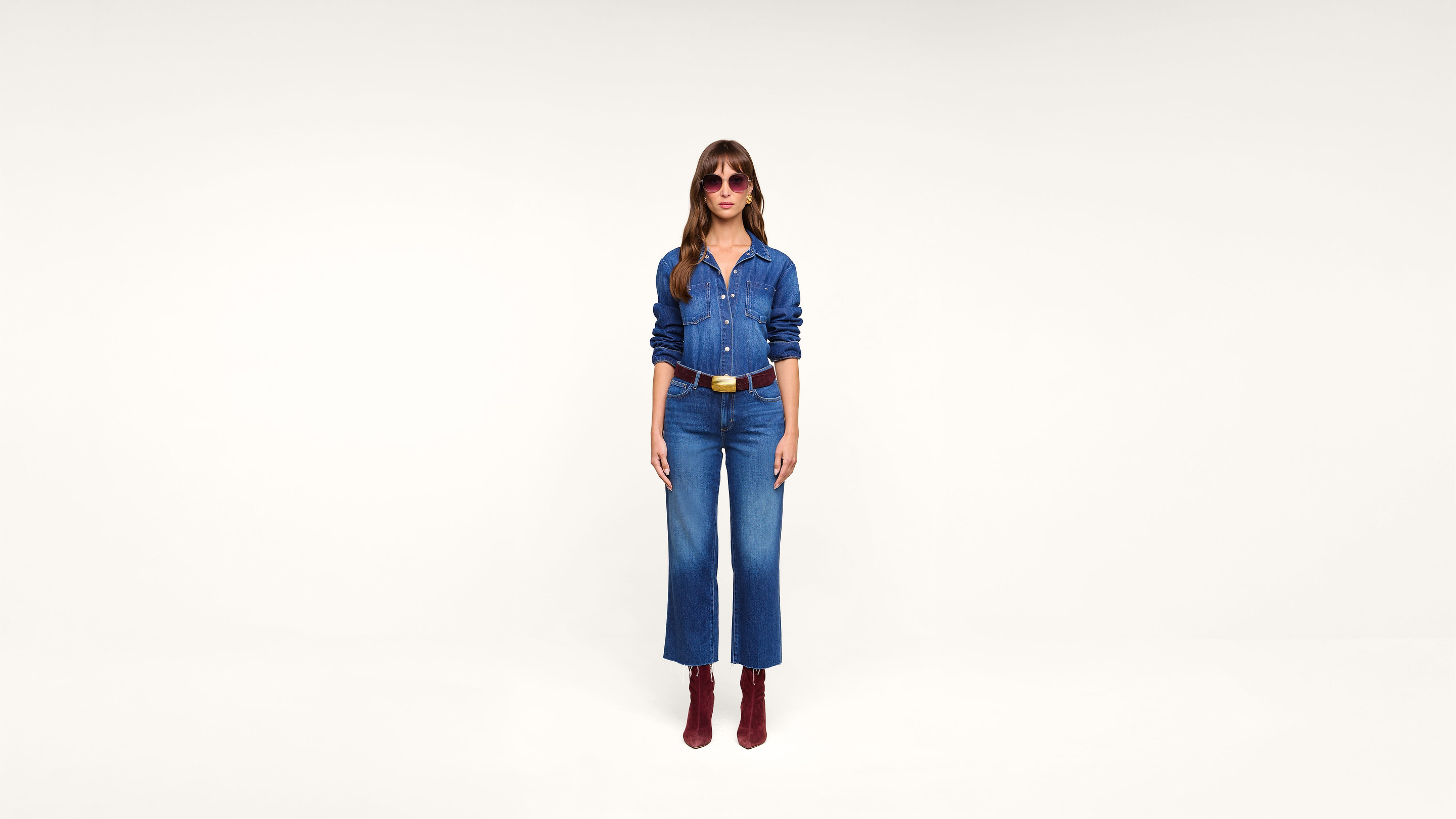 Woman wearing pink sunglasses, a denim shirt, blue jeans, brown belt, and red boots stands against a plain white background.