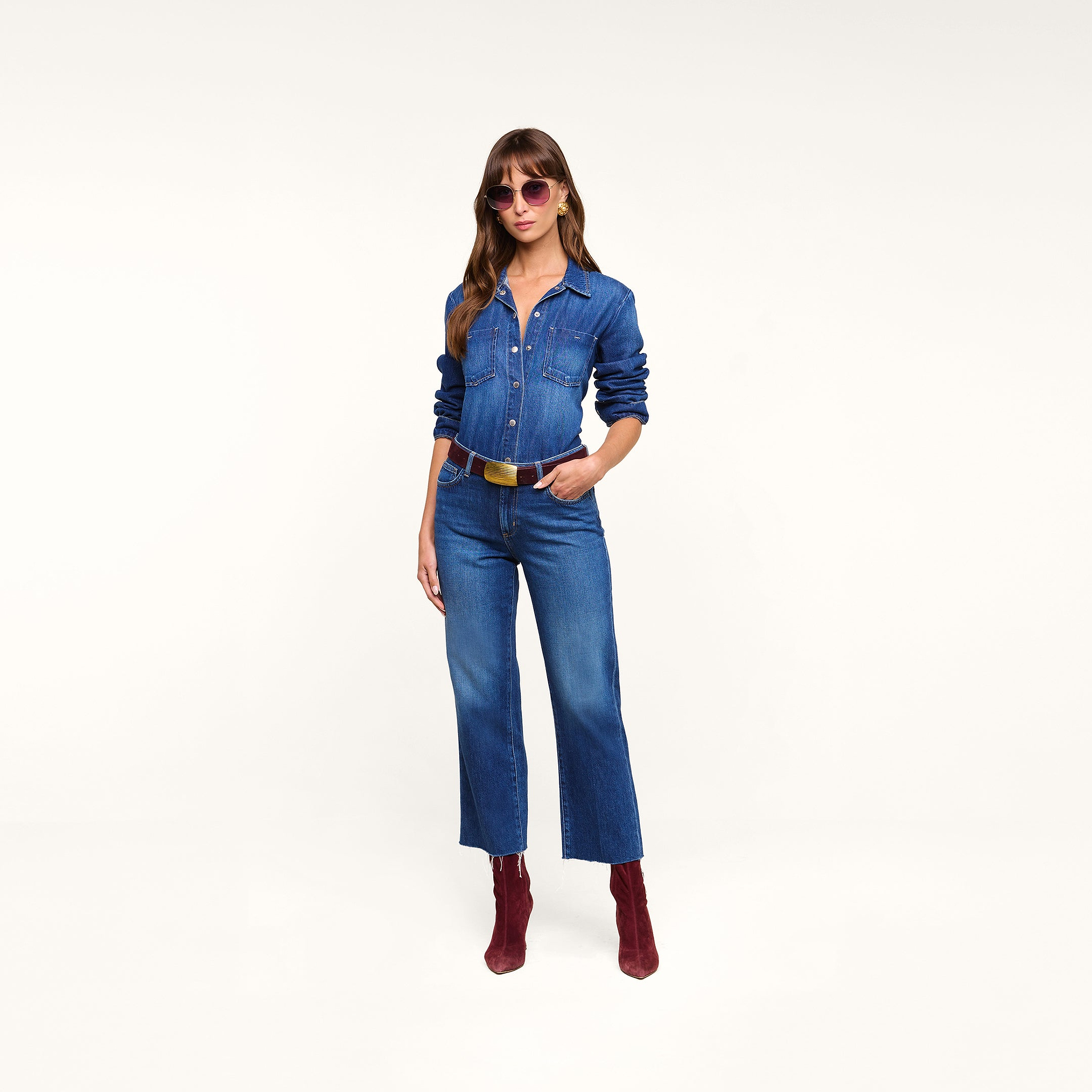 A woman stands with one hand in her pocket against a plain white background, wearing the Wanda Cropped Wide-Leg Jean, paired with red boots, sunglasses, and a black belt.
