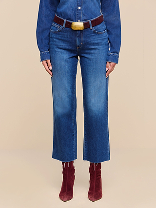Wearing the Wanda Cropped Wide-Leg Jean in high-rise blue denim with a raw hem and wide-leg silhouette crafted from featherweight denim, paired with a matching denim shirt, maroon belt, and maroon ankle boots. The background is a plain beige color.