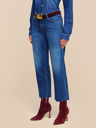 A person wearing the Wanda Cropped Wide-Leg Jean in high rise blue with frayed hems, paired with a featherweight denim shirt, a burgundy belt, and burgundy heeled ankle boots, standing against a beige background.