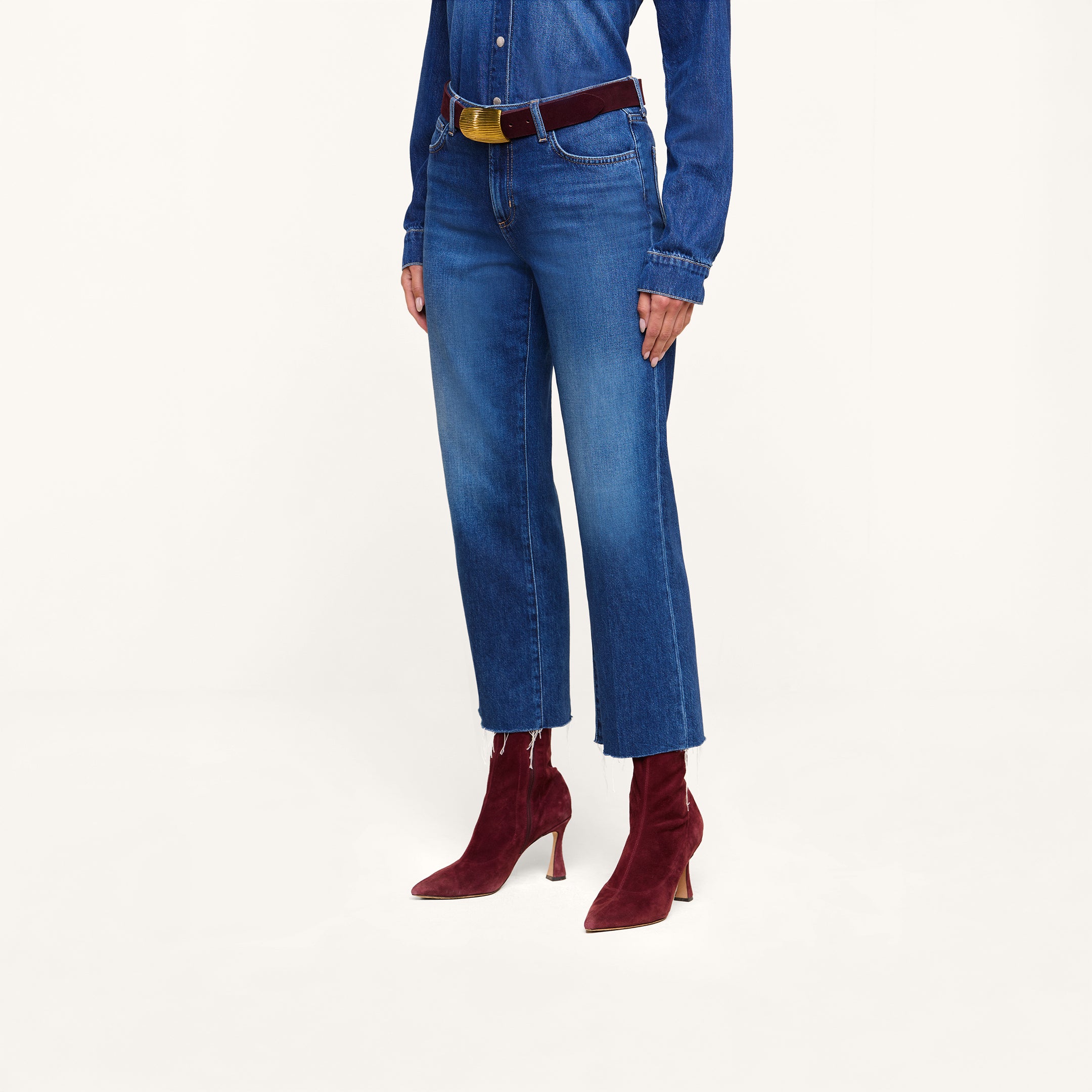 A person wearing the Wanda Cropped Wide-Leg Jean in featherweight denim, styled with a blue denim shirt, a dark belt, and pointed red suede ankle boots with heels, stands against a plain white background.
