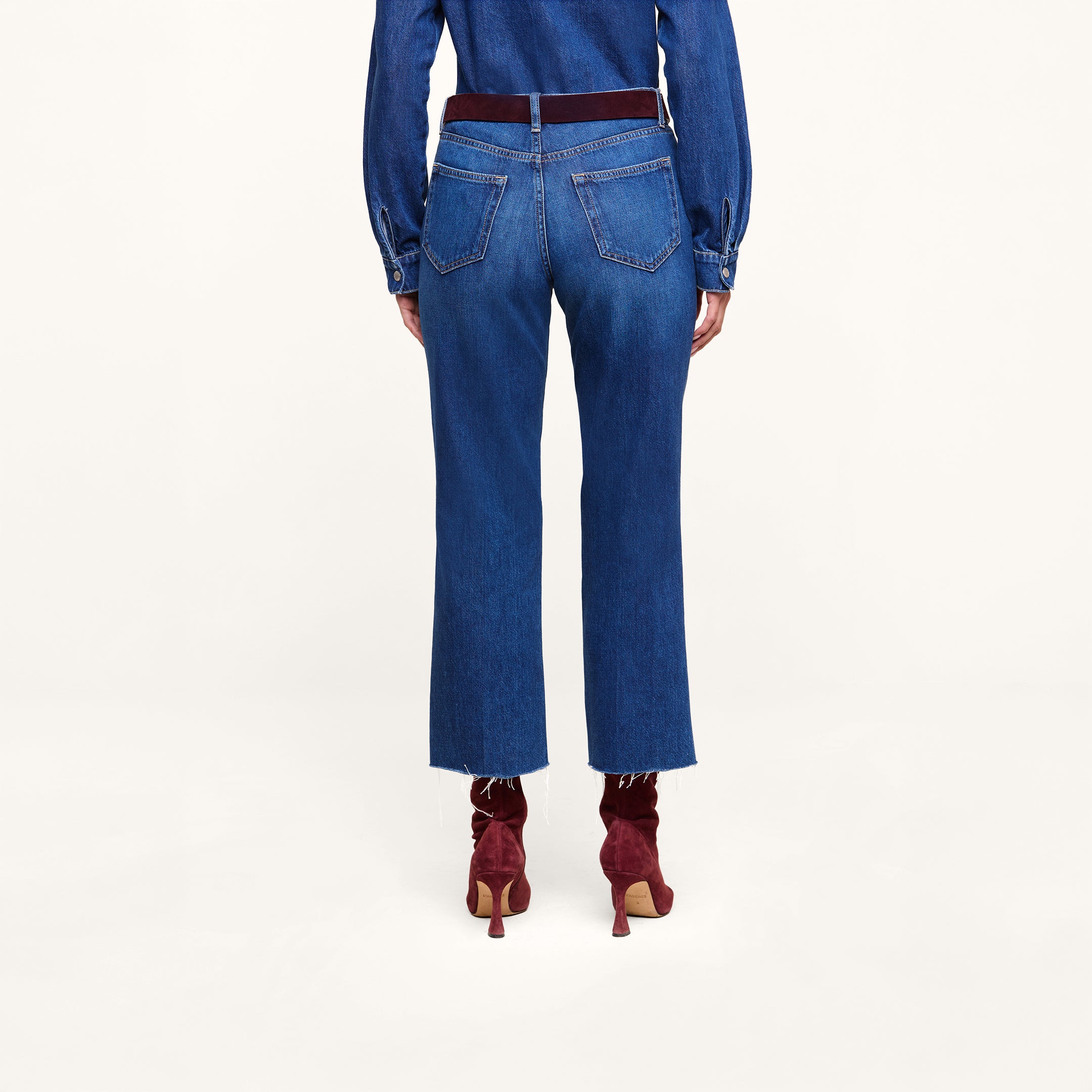 A person standing facing away on a plain white background wears a long-sleeve featherweight denim shirt, the Wanda Cropped Wide-Leg Jean featuring high rise styling and back pockets, accented with a dark red belt and matching dark red heeled ankle boots.