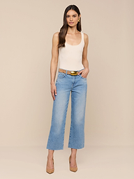 Eine Frau mit langen braunen Haaren posiert vor einem beigen Hintergrund. Sie trägt ein cremefarbenes Tank-Top und eine hellblaue Wanda Cropped Wide-Leg Jeans, die mit einem hellbraunen Gürtel und hellbraunen High Heels gestylt ist. Eine Hand ruht in ihrer Tasche.