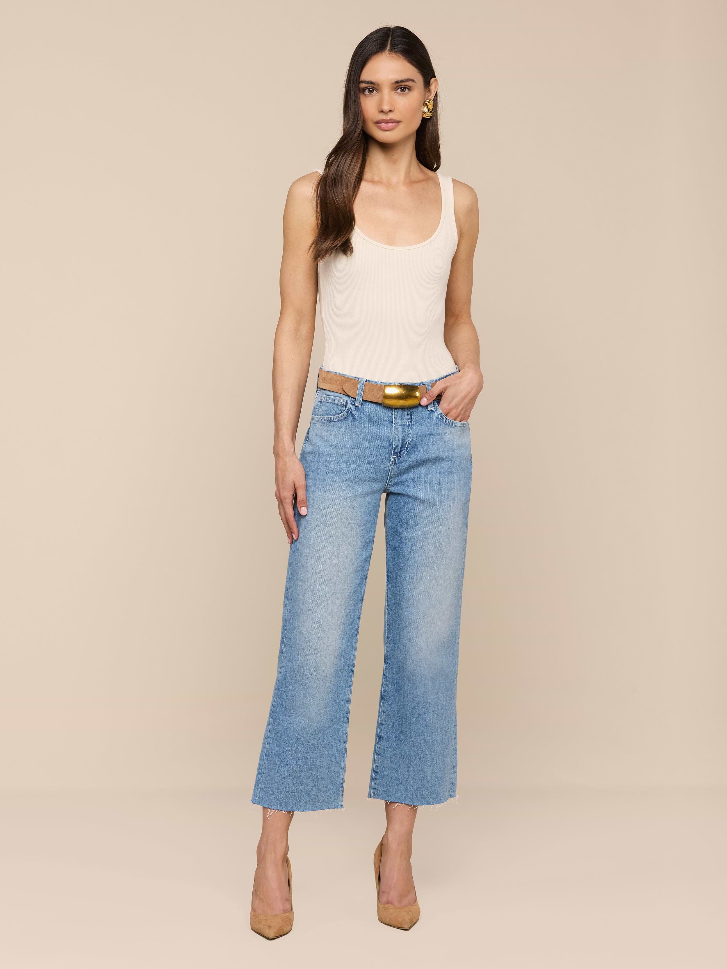 Eine Frau mit langen braunen Haaren posiert vor einem beigen Hintergrund. Sie trägt ein cremefarbenes Tank-Top und eine hellblaue Wanda Cropped Wide-Leg Jeans, die mit einem hellbraunen Gürtel und hellbraunen High Heels gestylt ist. Eine Hand ruht in ihrer Tasche.